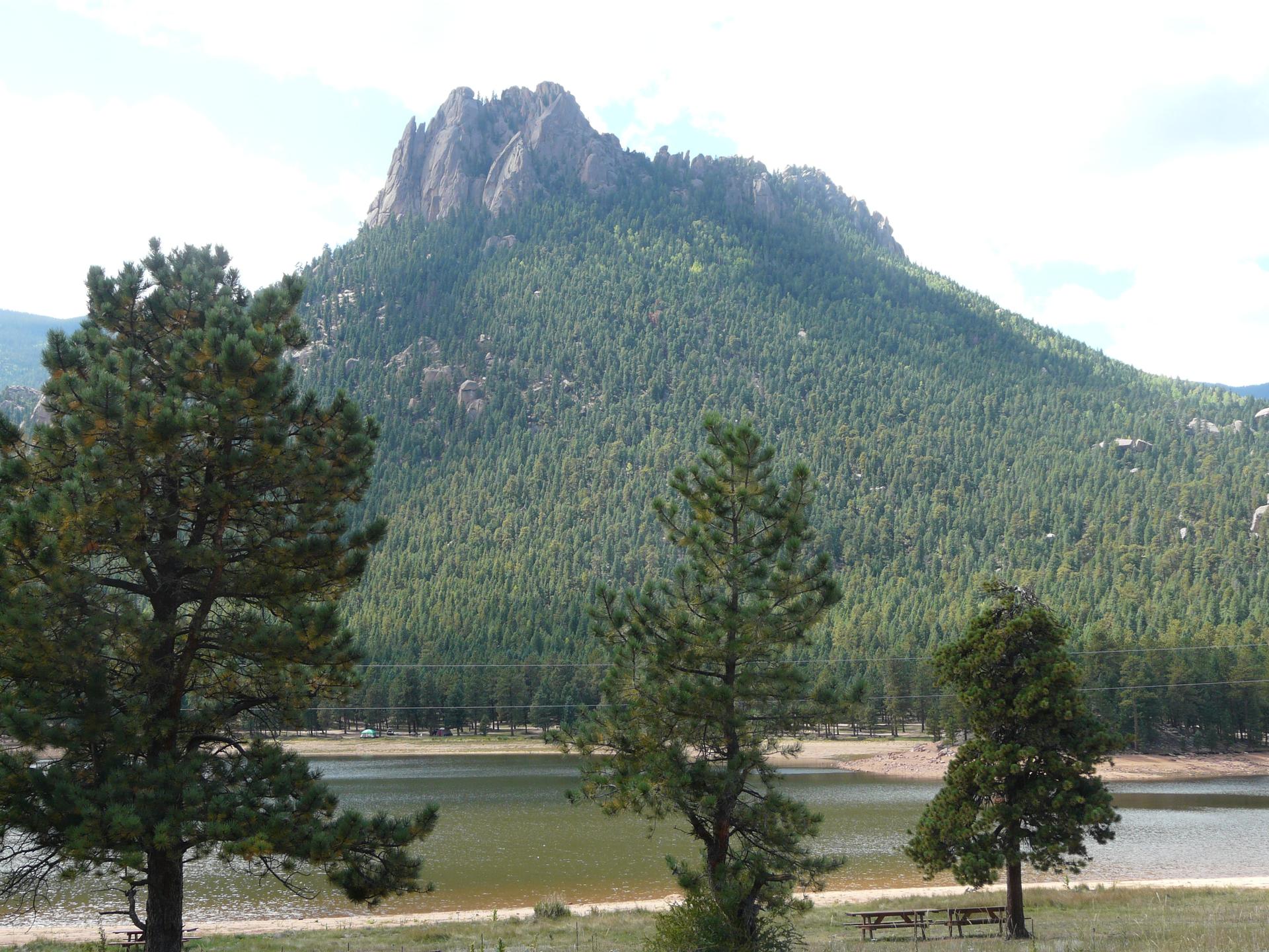 Sunny view of a rocky, forested mountain overlooking a lake with pine trees.