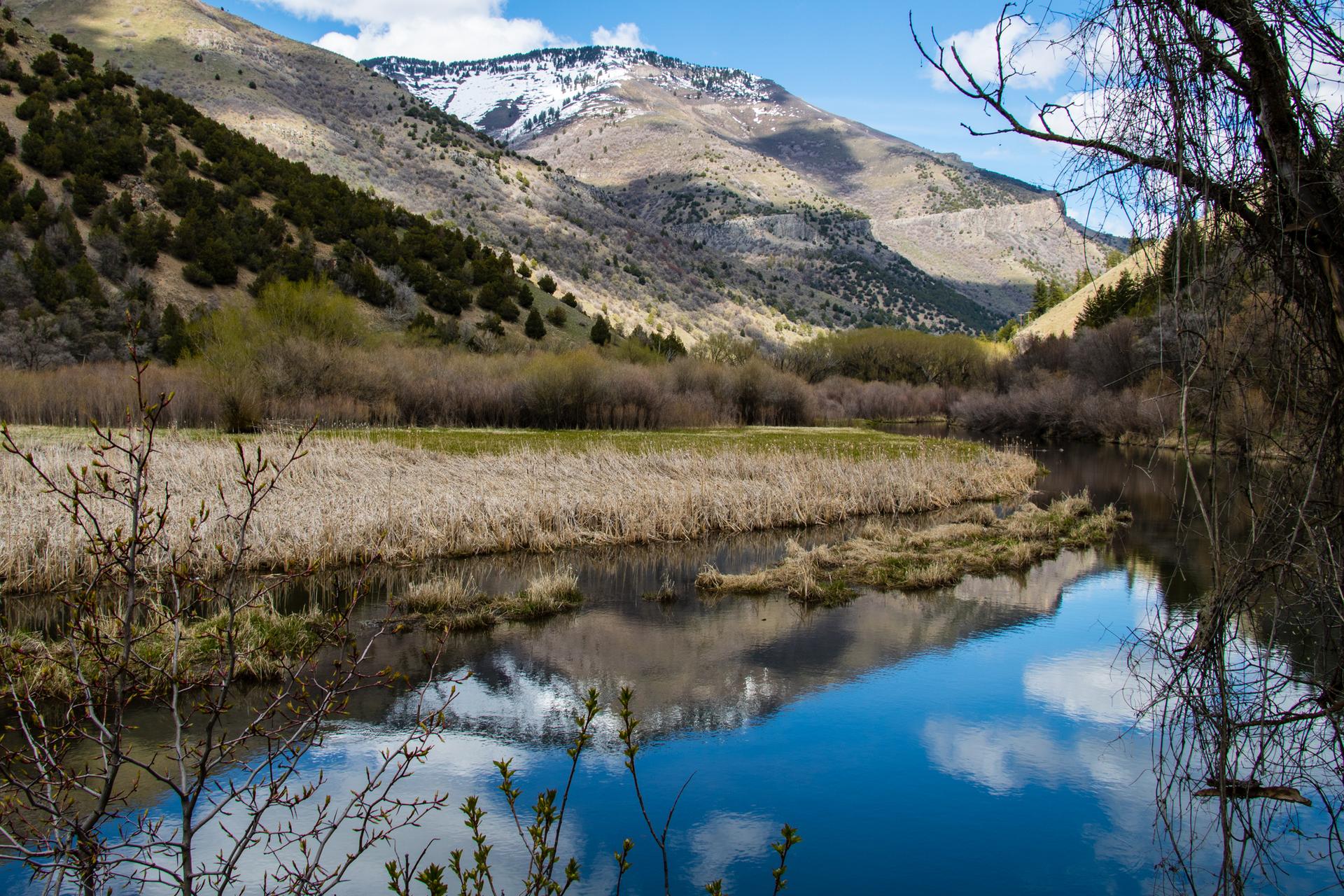 A calm river reflects snow-capped mountains and a blue sky with clouds.