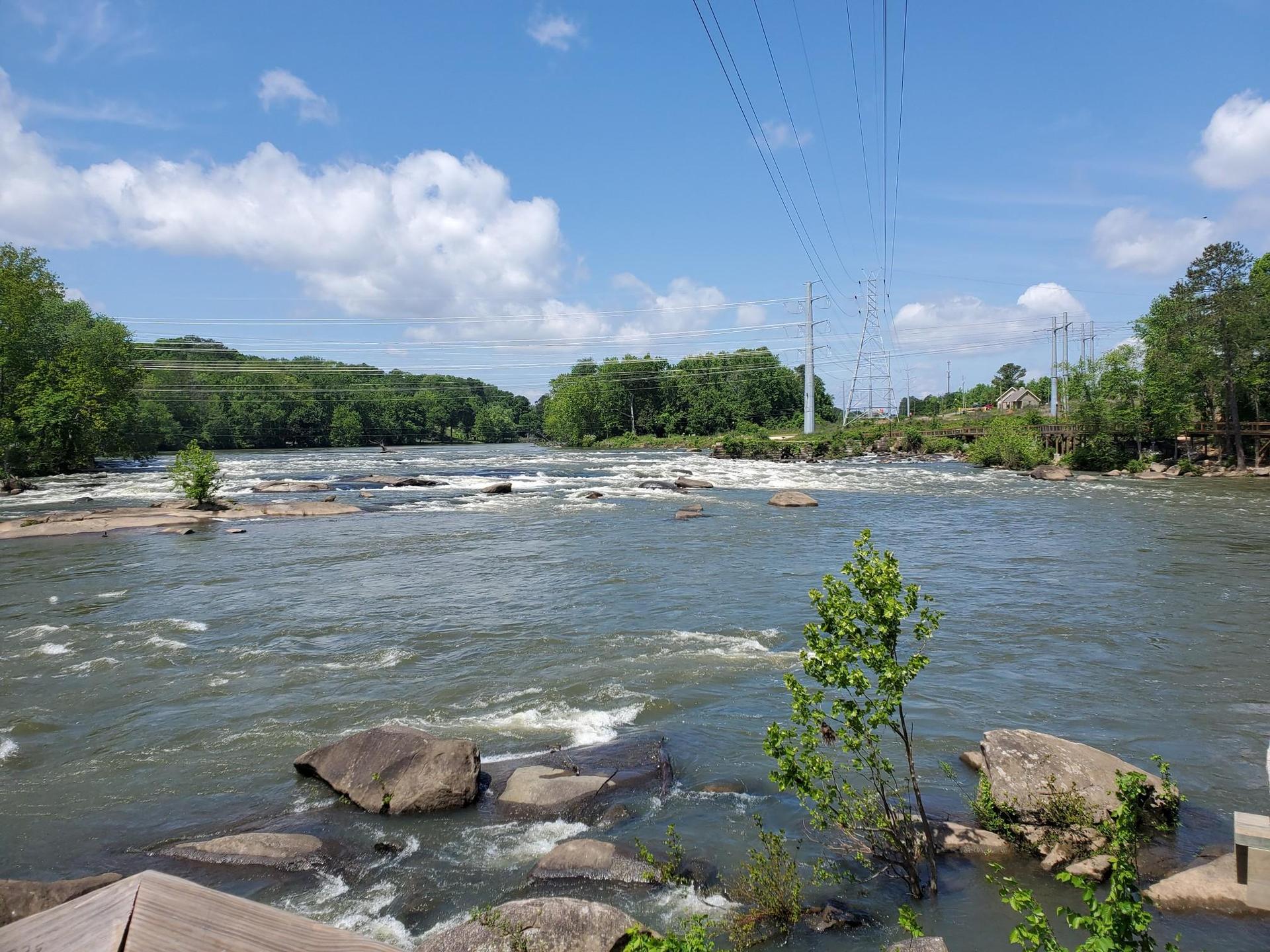 Wide river with rapids, green trees, and power lines under a blue sky.