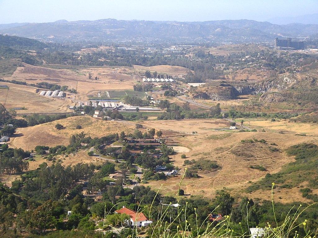 Expansive view of a dry valley with scattered buildings and distant hills under bright sun.