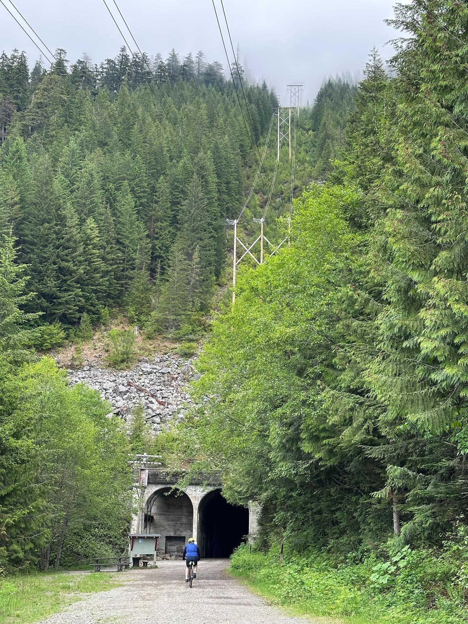Cyclist approaches two dark tunnel entrances on a gravel path in a green forest.