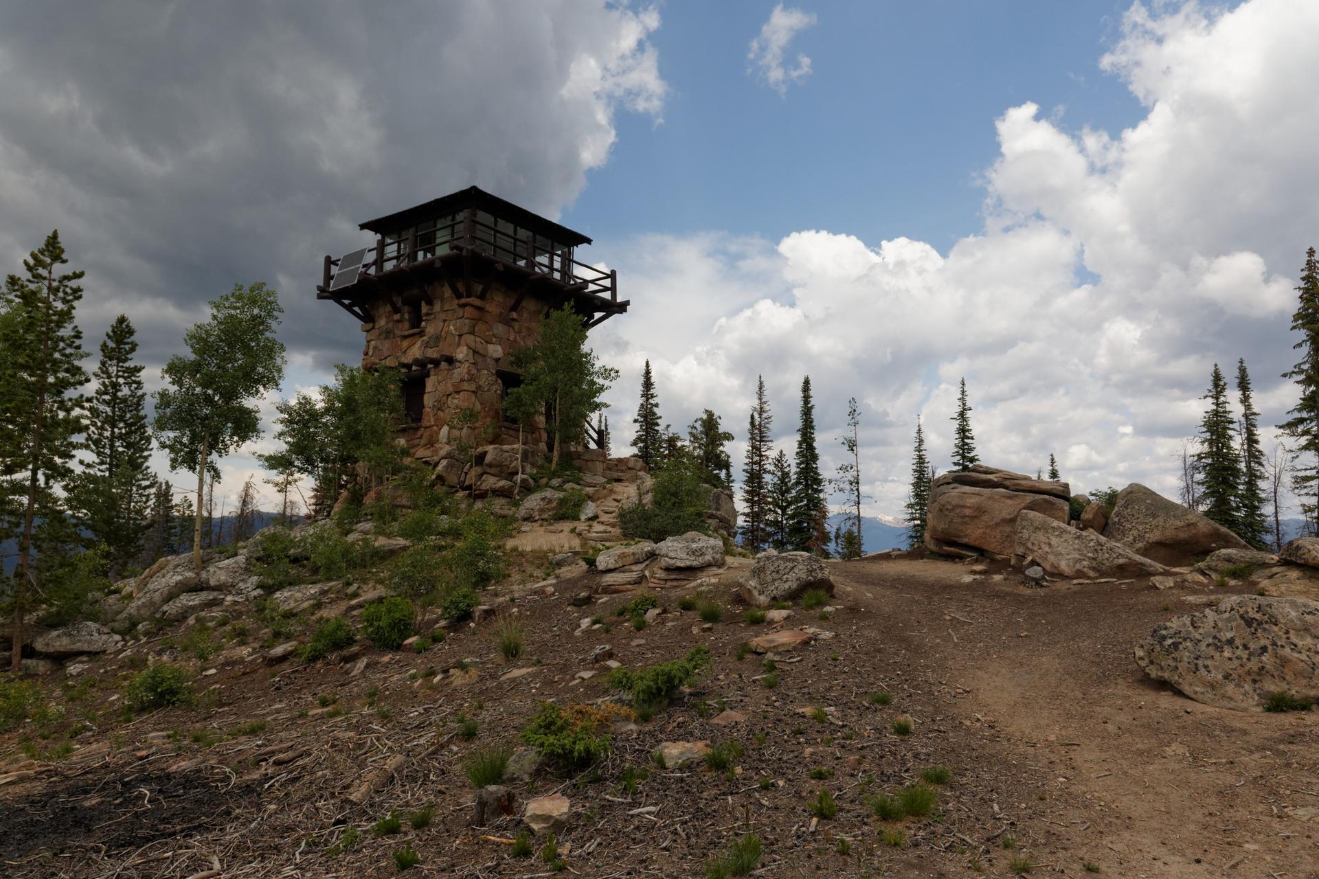A stone fire lookout tower sits atop a rocky hill under a cloudy sky.