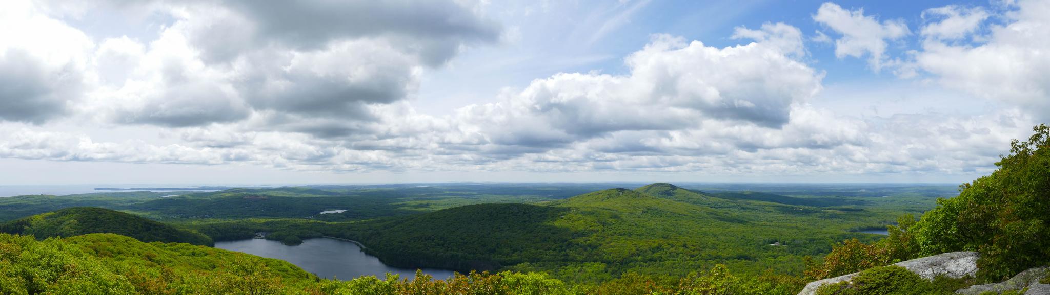 Panoramic view of lakes and vast green forested hills under a partly cloudy sky.