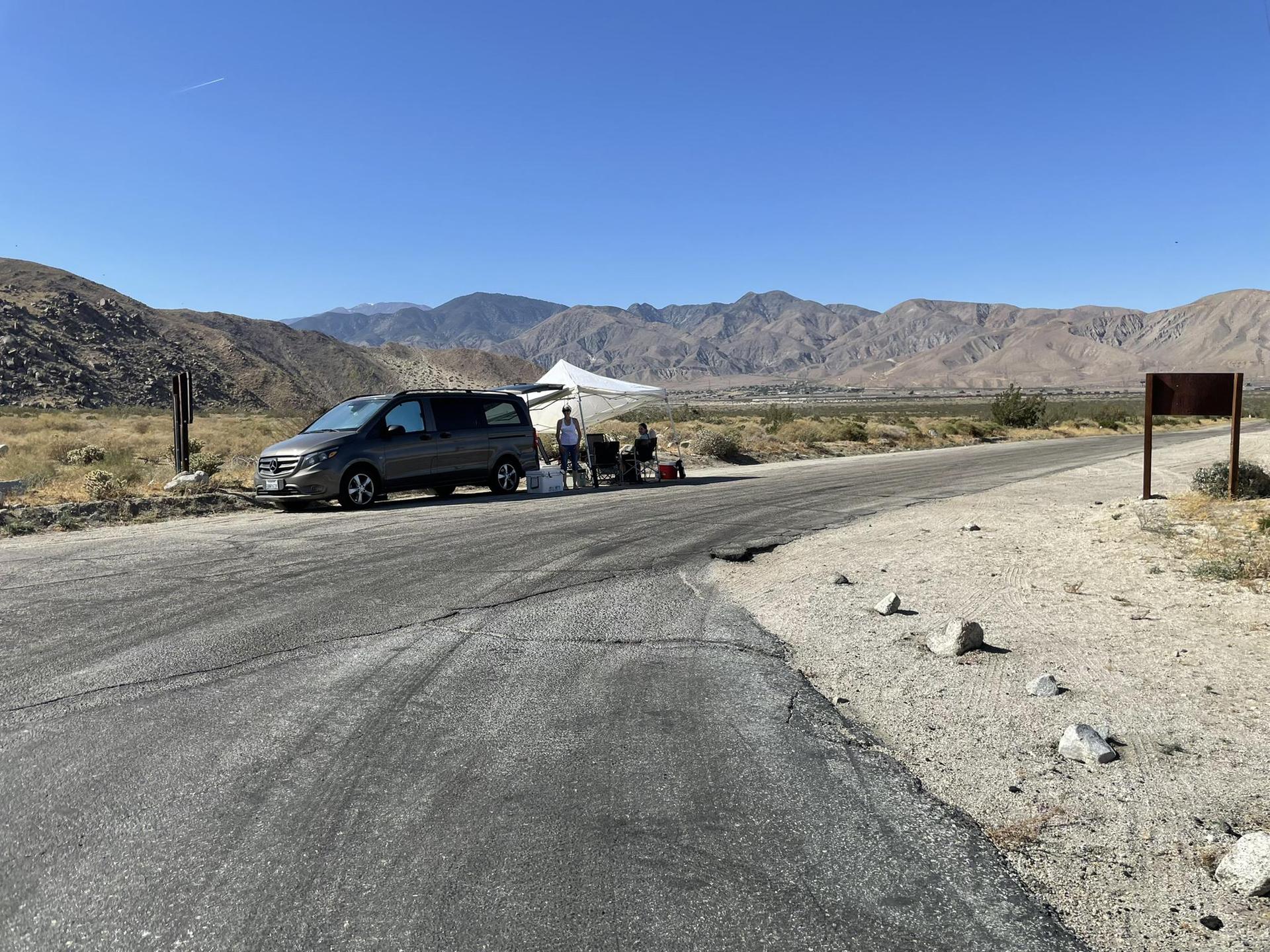 Van and people under a white canopy beside a road in a desert.