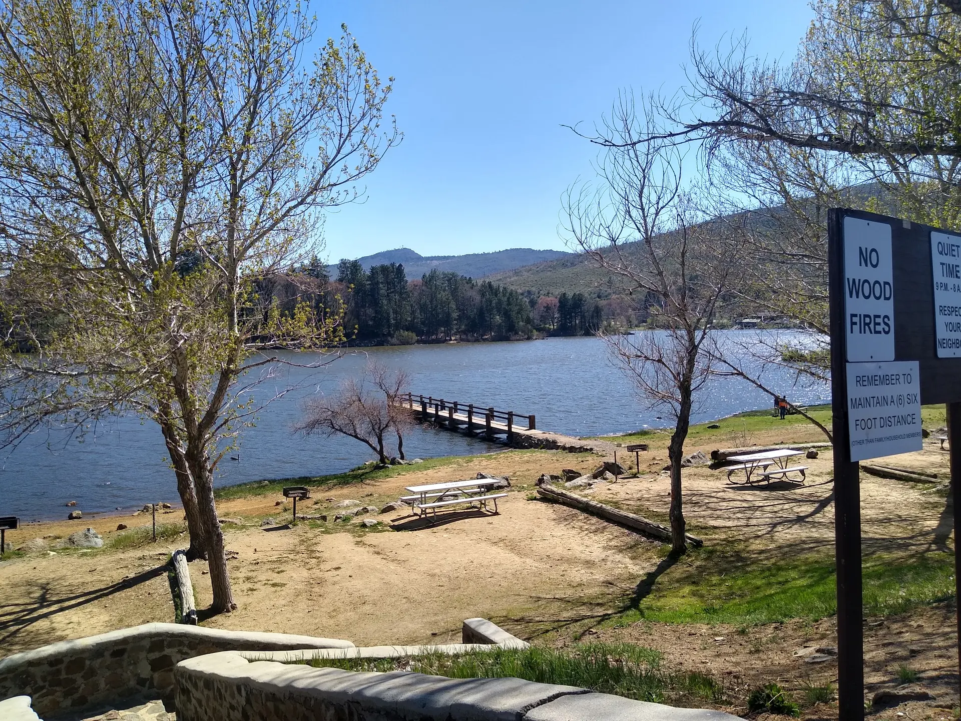 Sunny lakeside picnic area with a dock, trees, and distant mountains.