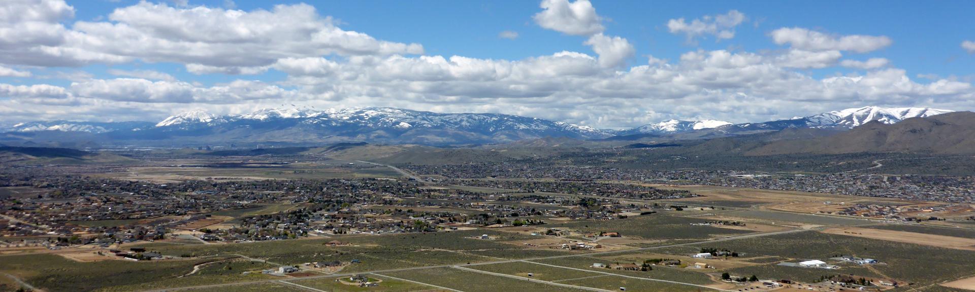 Expansive valley with a town, green fields, and snowy mountains under blue sky.