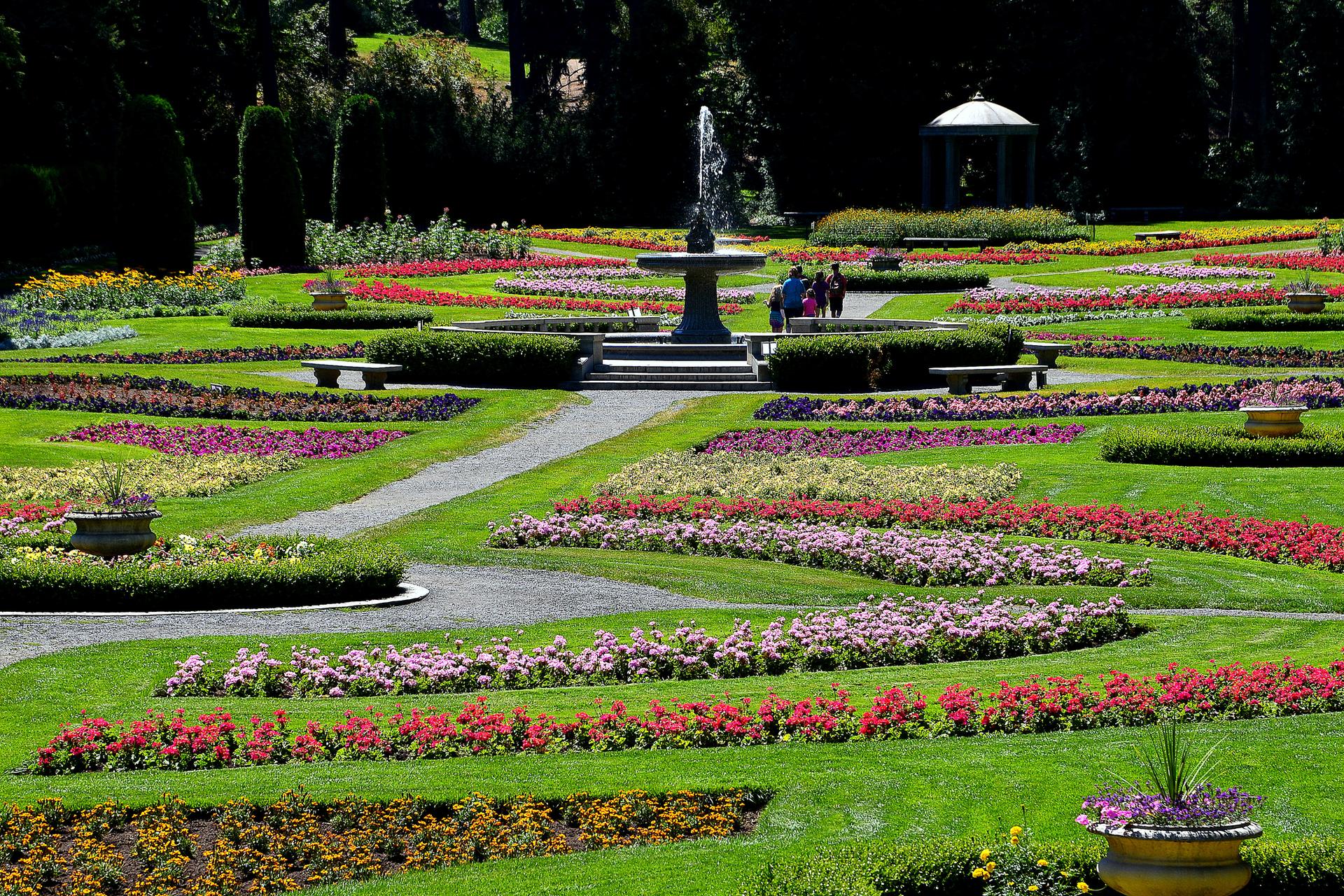Vibrant formal garden with a central fountain, colorful flowerbeds, and distant people.