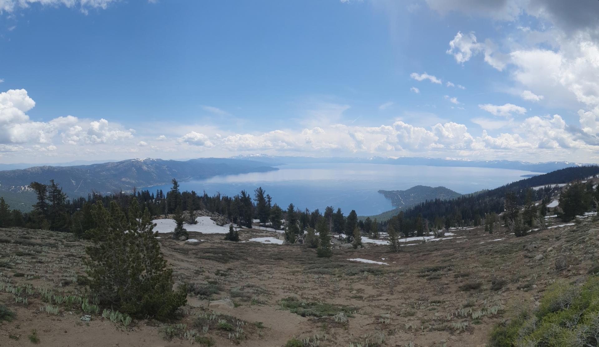 Panoramic view of a vast lake surrounded by mountains, trees, and snow patches.