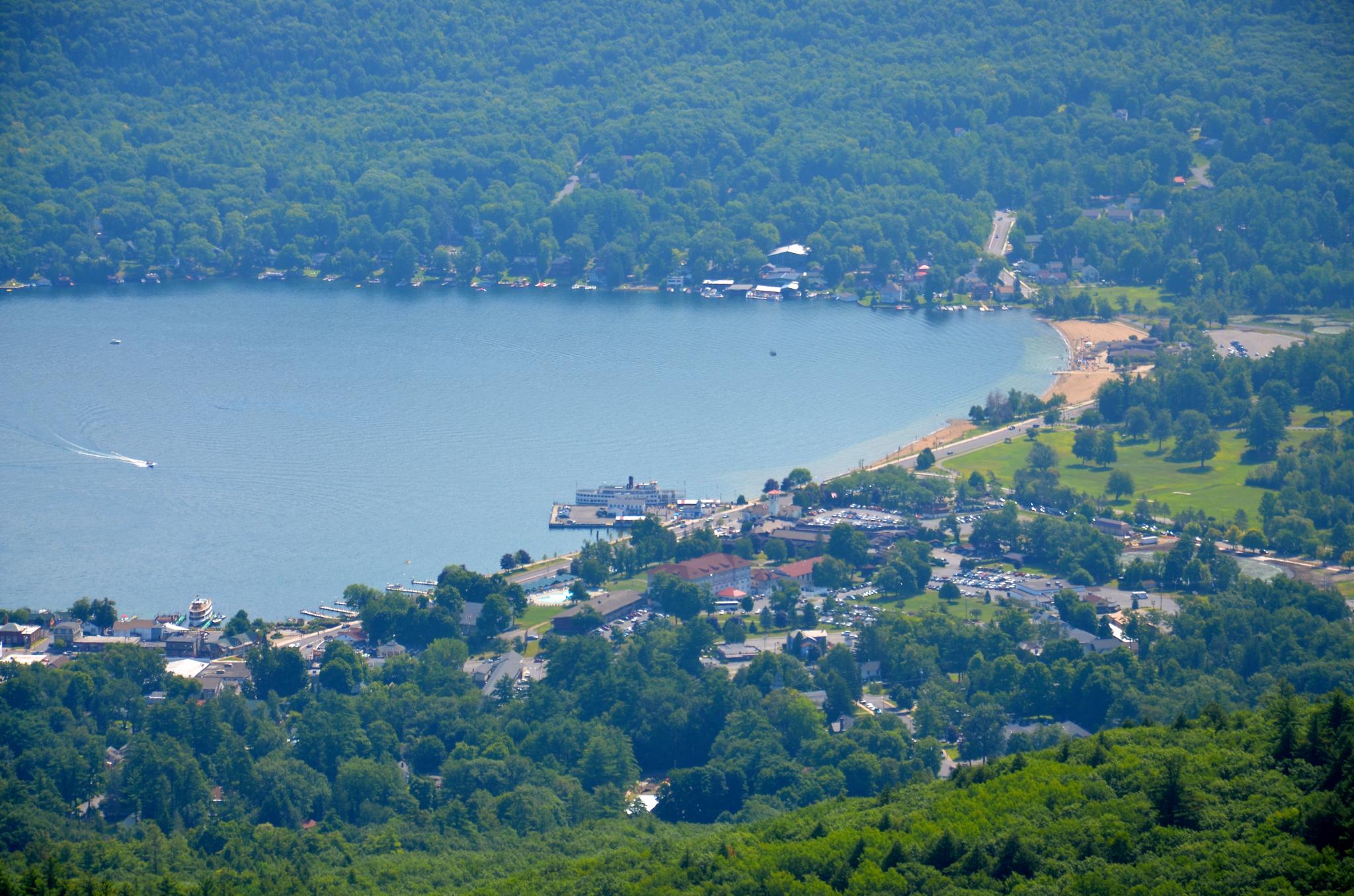 Aerial view of a lake with a town, beach, and boats under a clear sky.