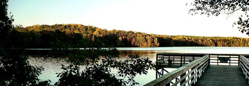 Wooden dock extending into a calm lake with autumn trees at sunset.