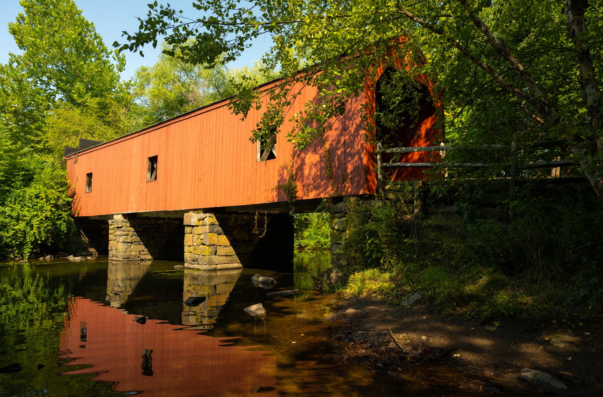 Red covered bridge over a calm river, surrounded by lush green trees.
