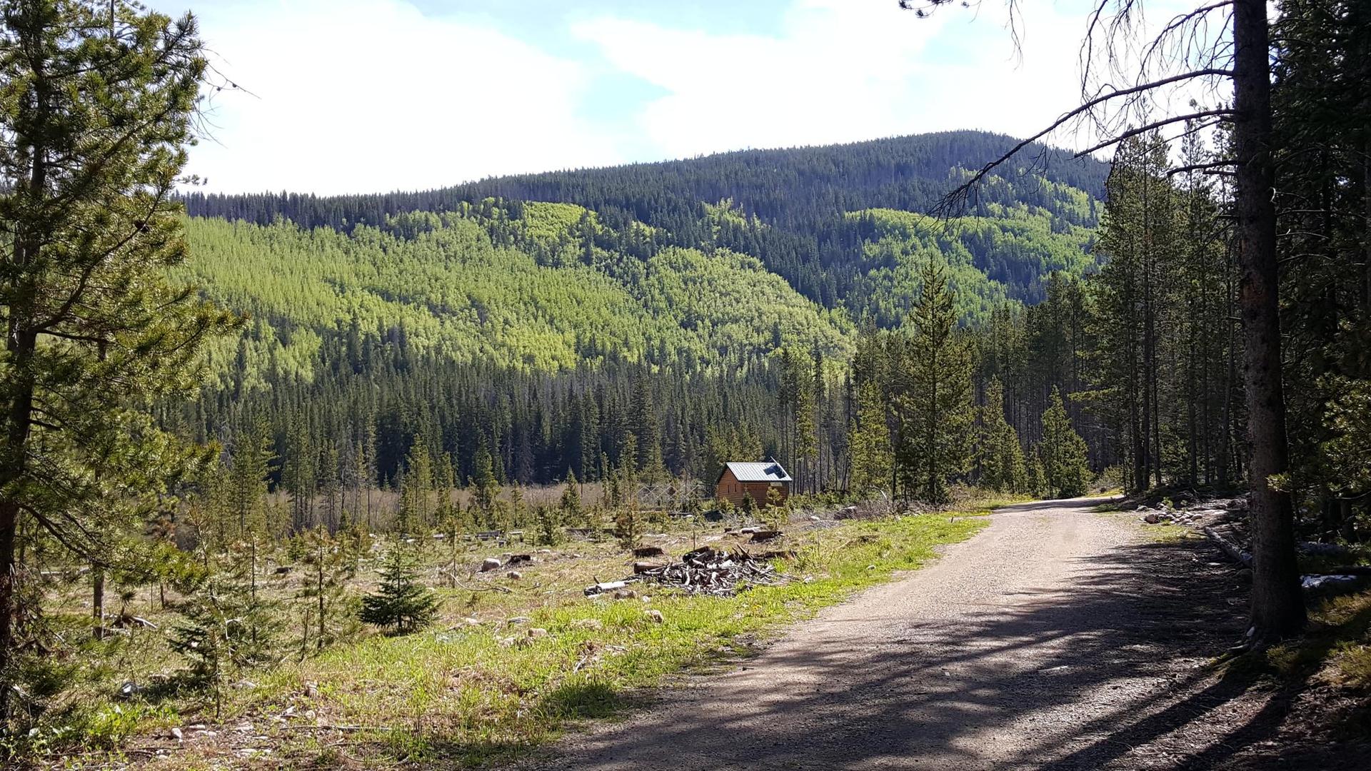 A small cabin sits in a clearing below a forested mountain.