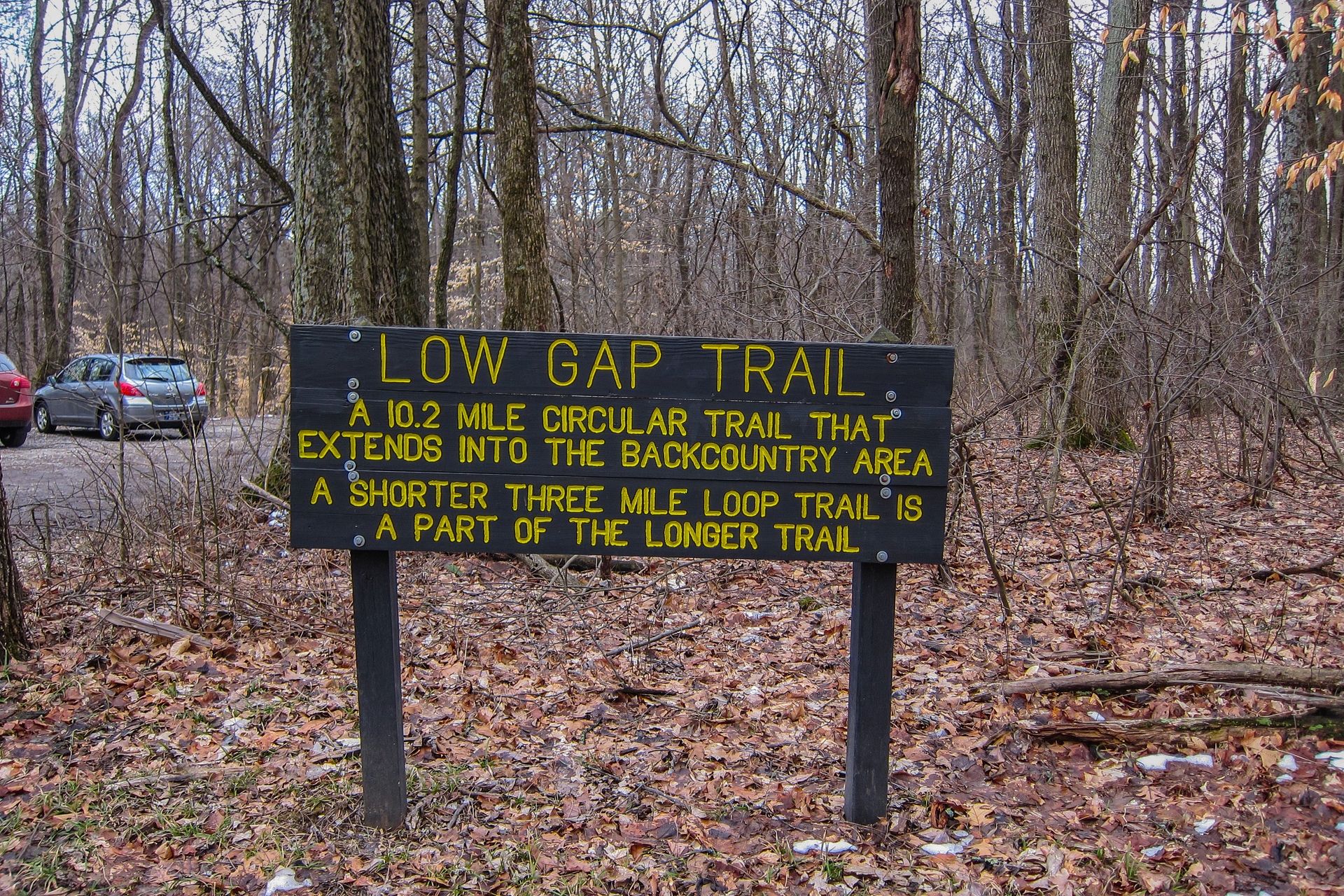 A dark wooden sign with yellow text stands in a winter forest with parked cars. - Mason Ridge Trail