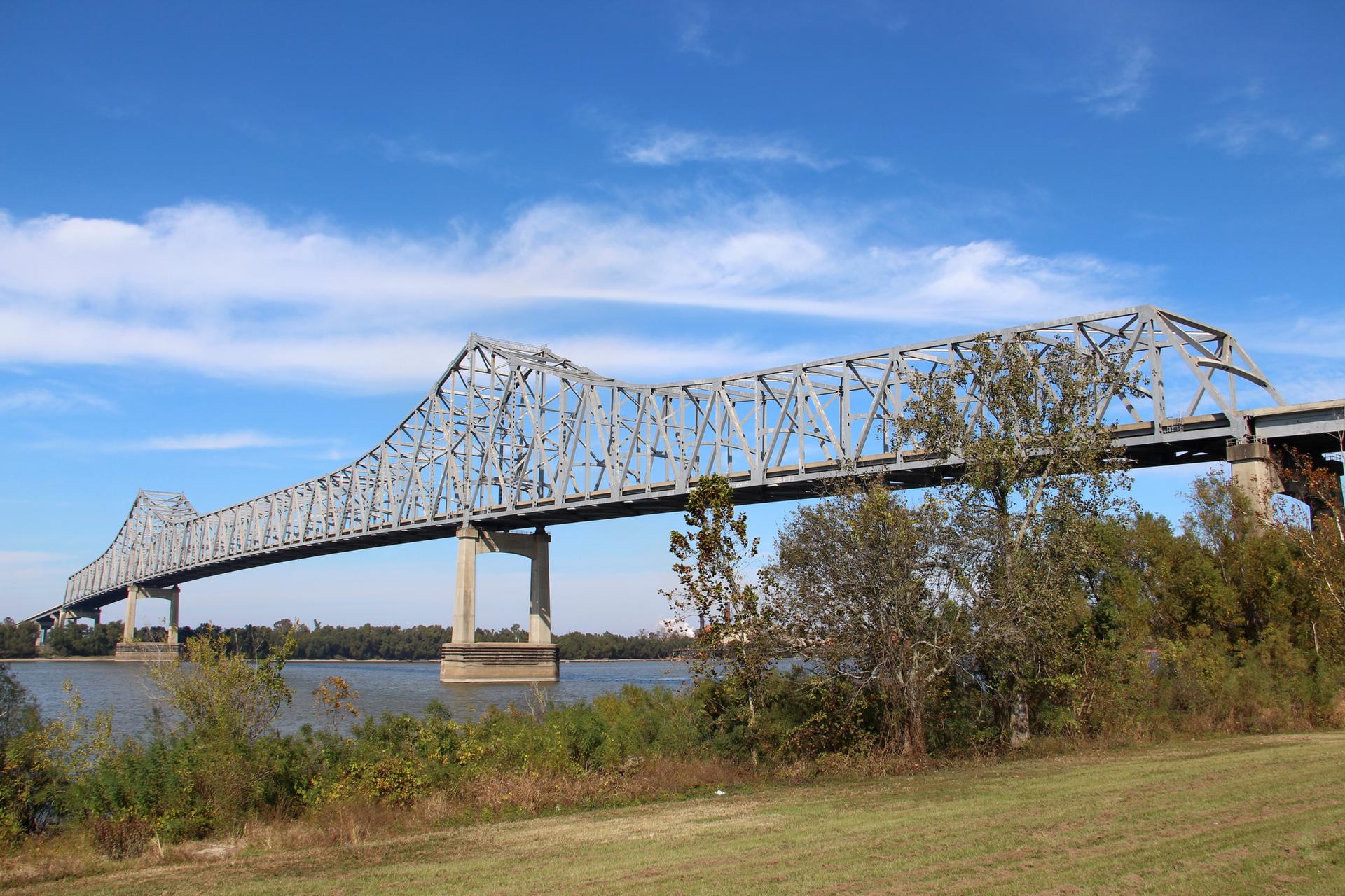 A large metal truss bridge spans a wide river under a blue sky.