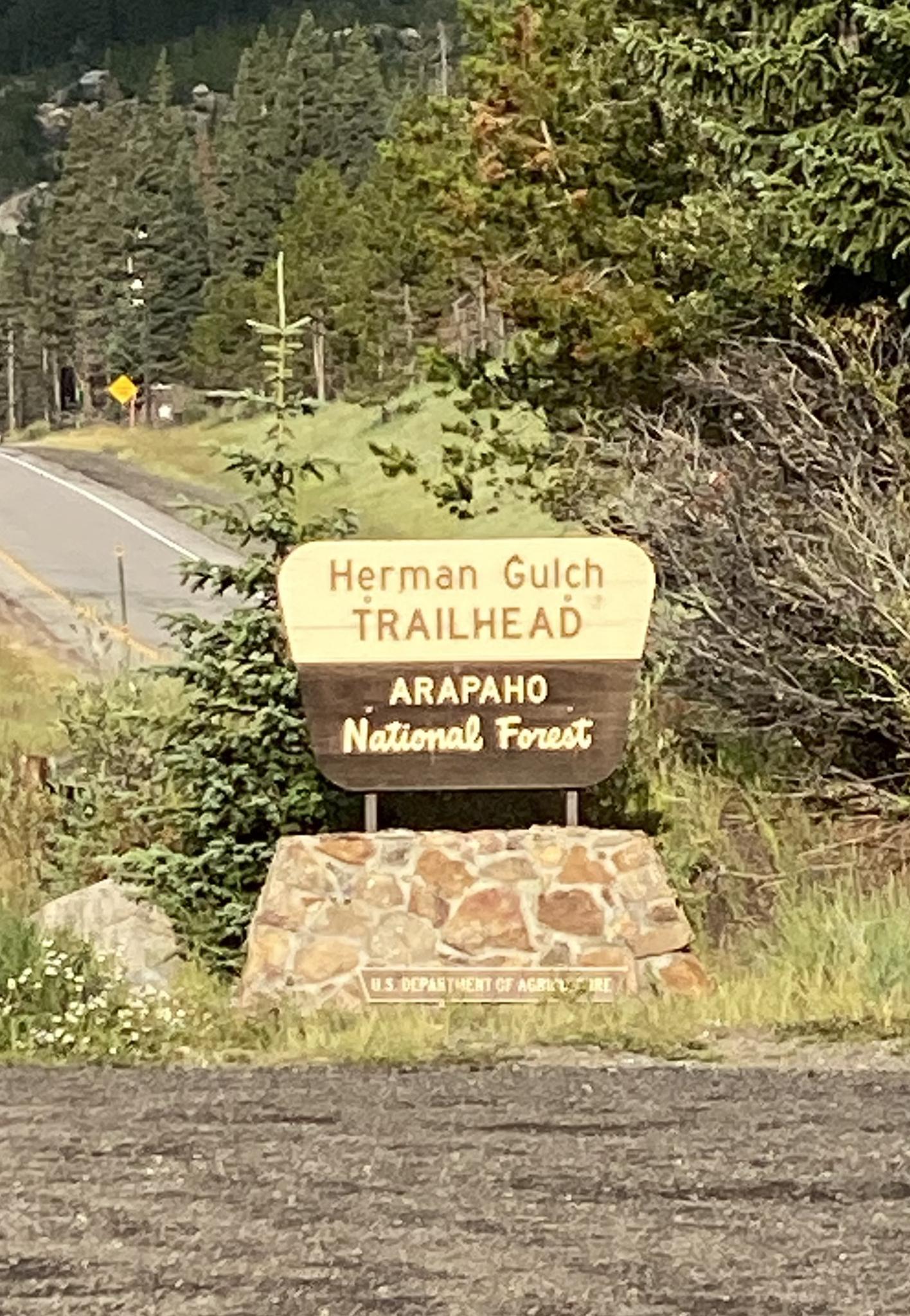 A wooden trailhead sign on a stone base next to a paved road.