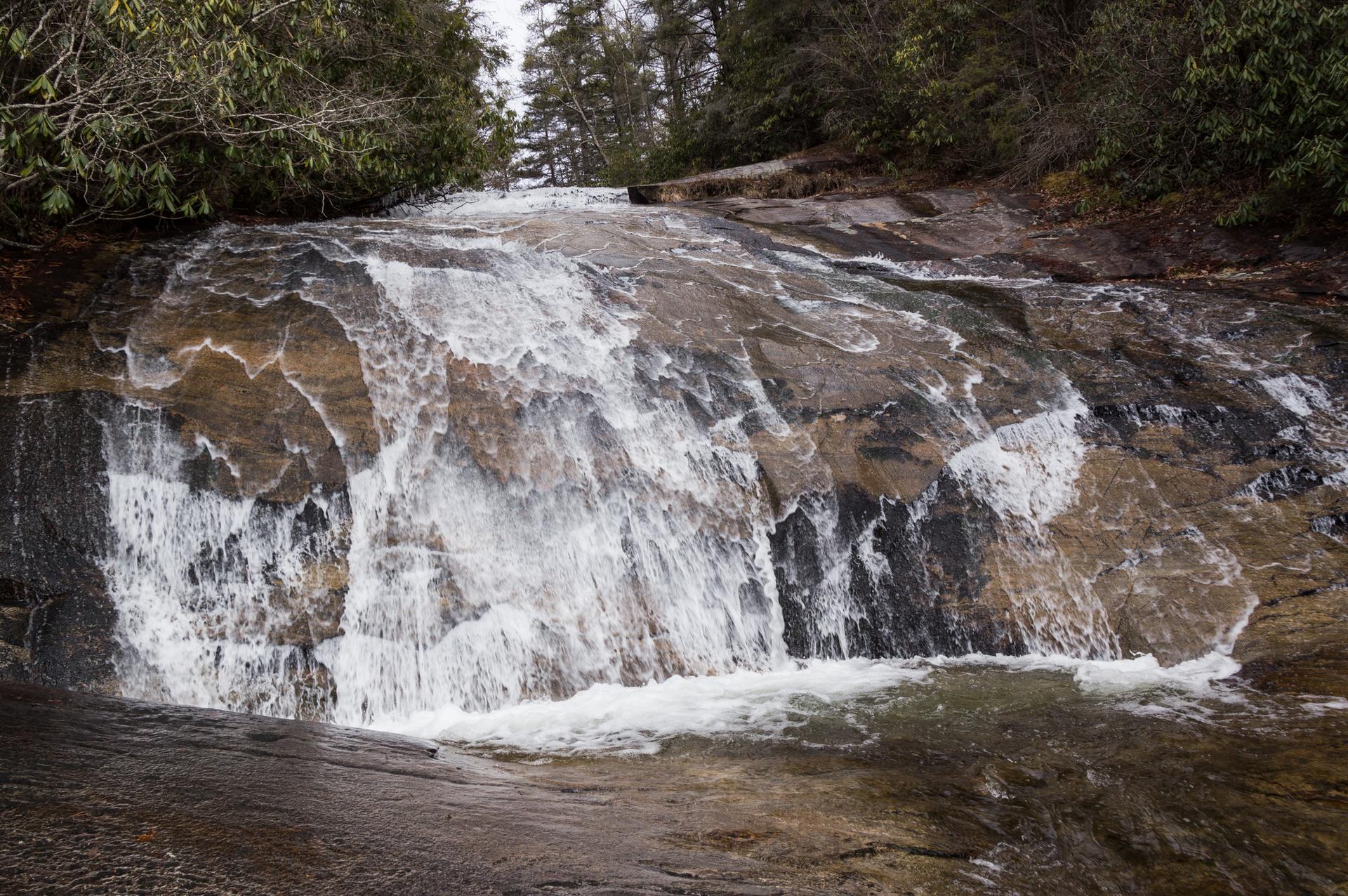 Waterfall cascades over dark, wet rocks with trees in the background.