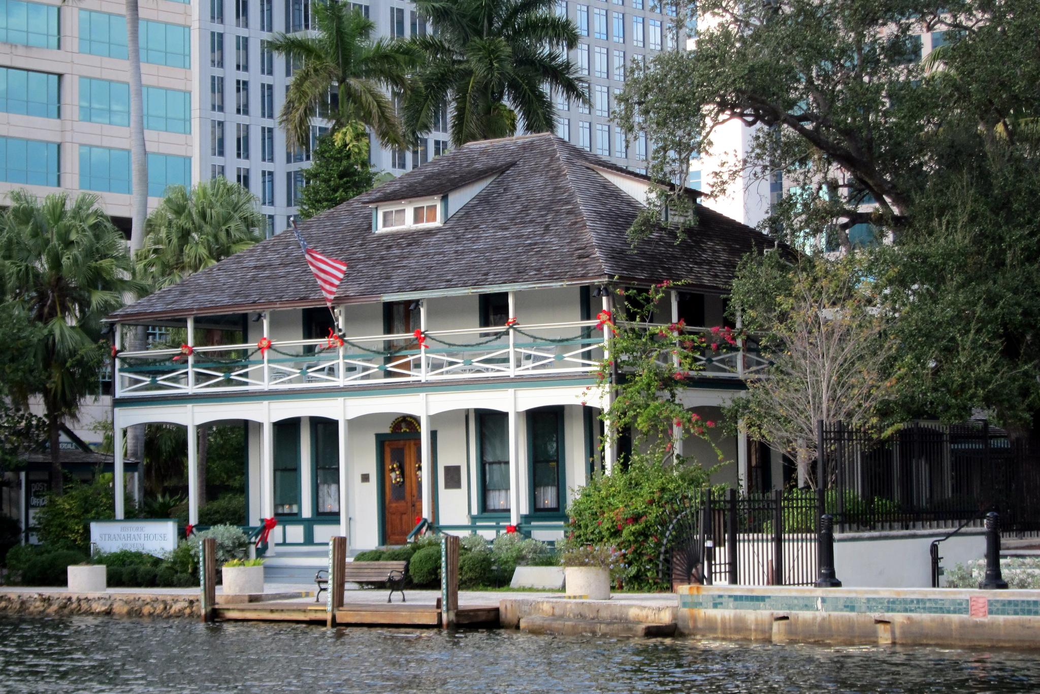 Historic house with balconies by water, modern buildings in the background.