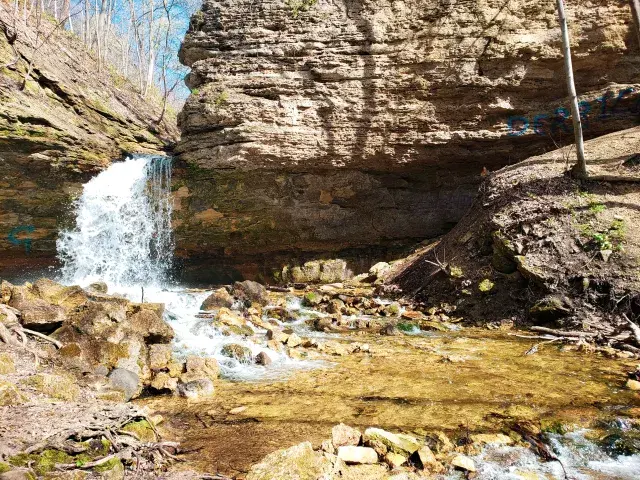 Waterfall cascades over rocky cliffs into a shallow stream on a sunny day.