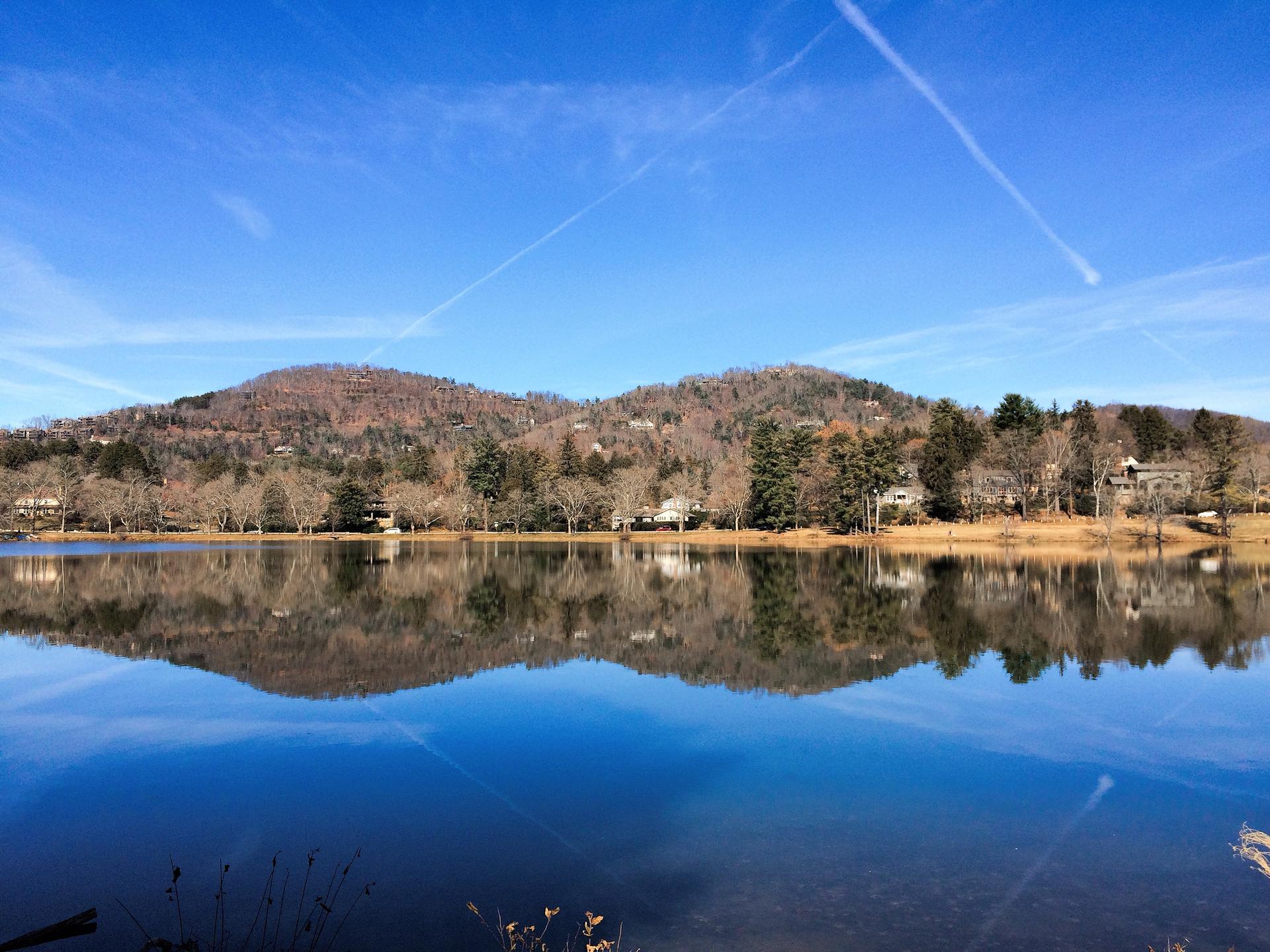 Calm lake reflecting tree-covered mountains under a bright blue sky.
