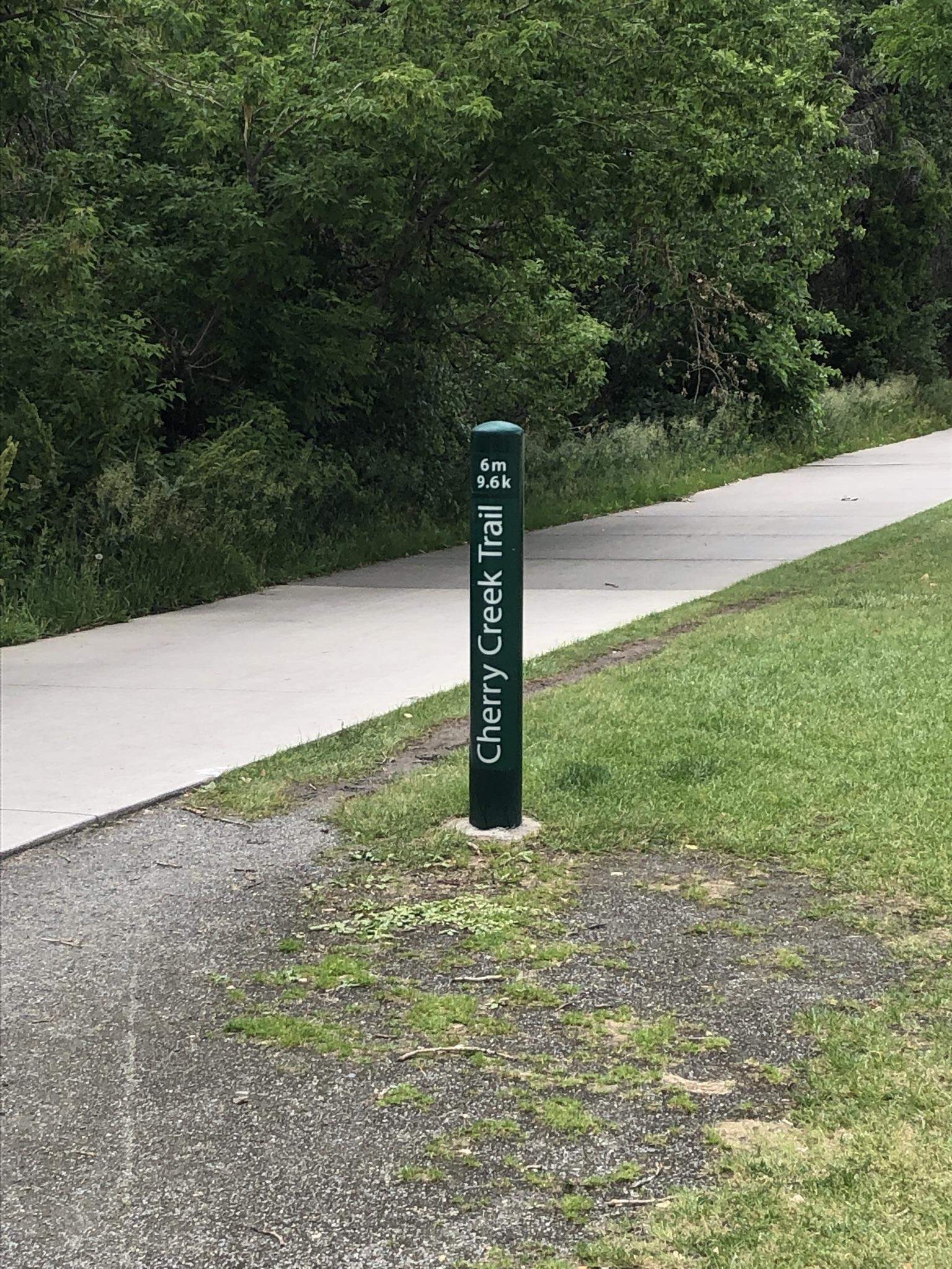 A green marker post stands beside a paved trail with green grass and trees.