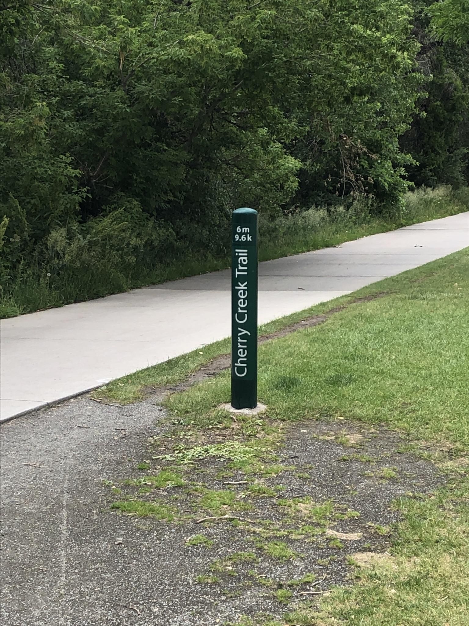A green marker post stands beside a paved trail with green grass and trees. - Four Mile Historic Park – Cherry Creek Trail North loop from Glendale