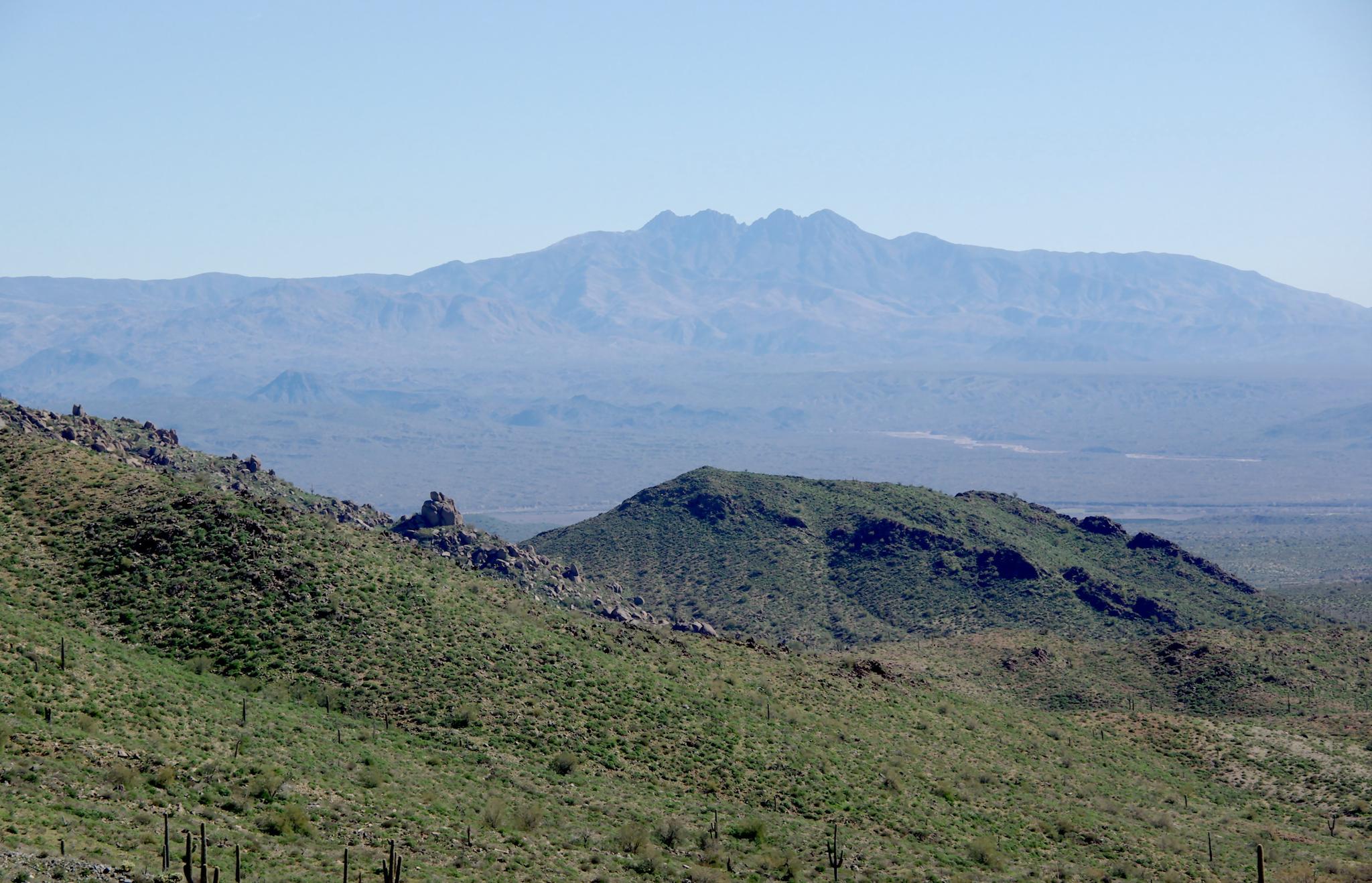 Sunny desert view with green, rocky hills and distant, hazy blue mountains.