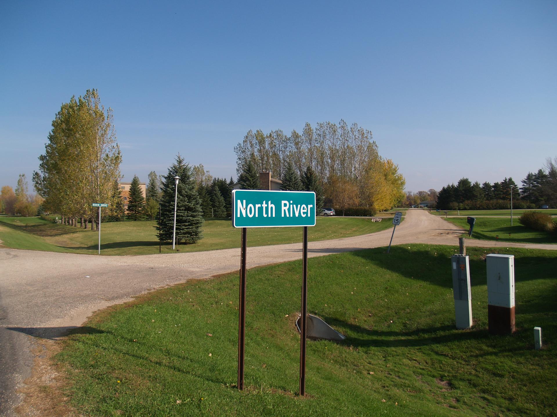 A green sign marks a dirt road junction, surrounded by trees and grass on a sunny day.