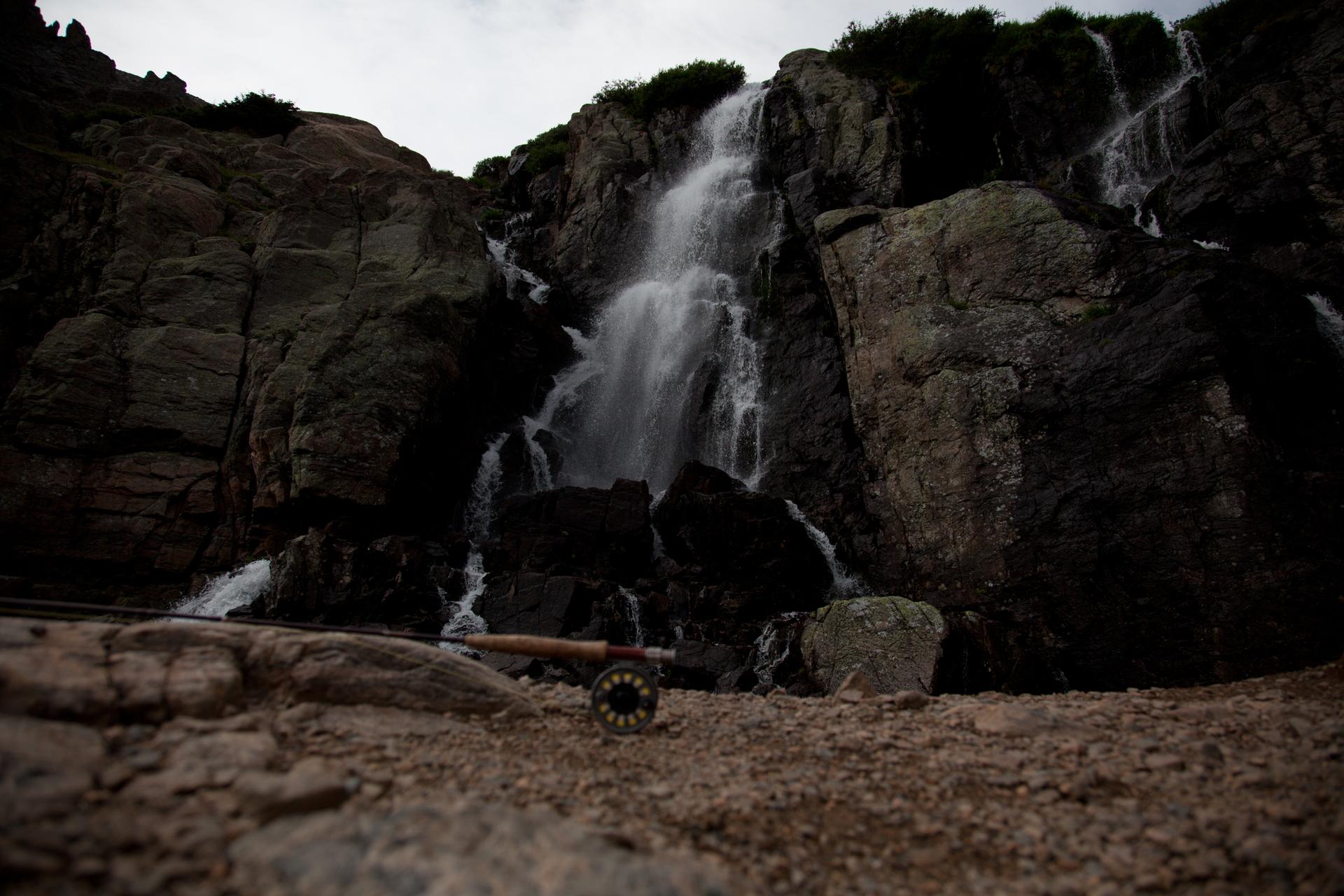 A fishing reel rests on rocky ground before a dark, cascading waterfall.