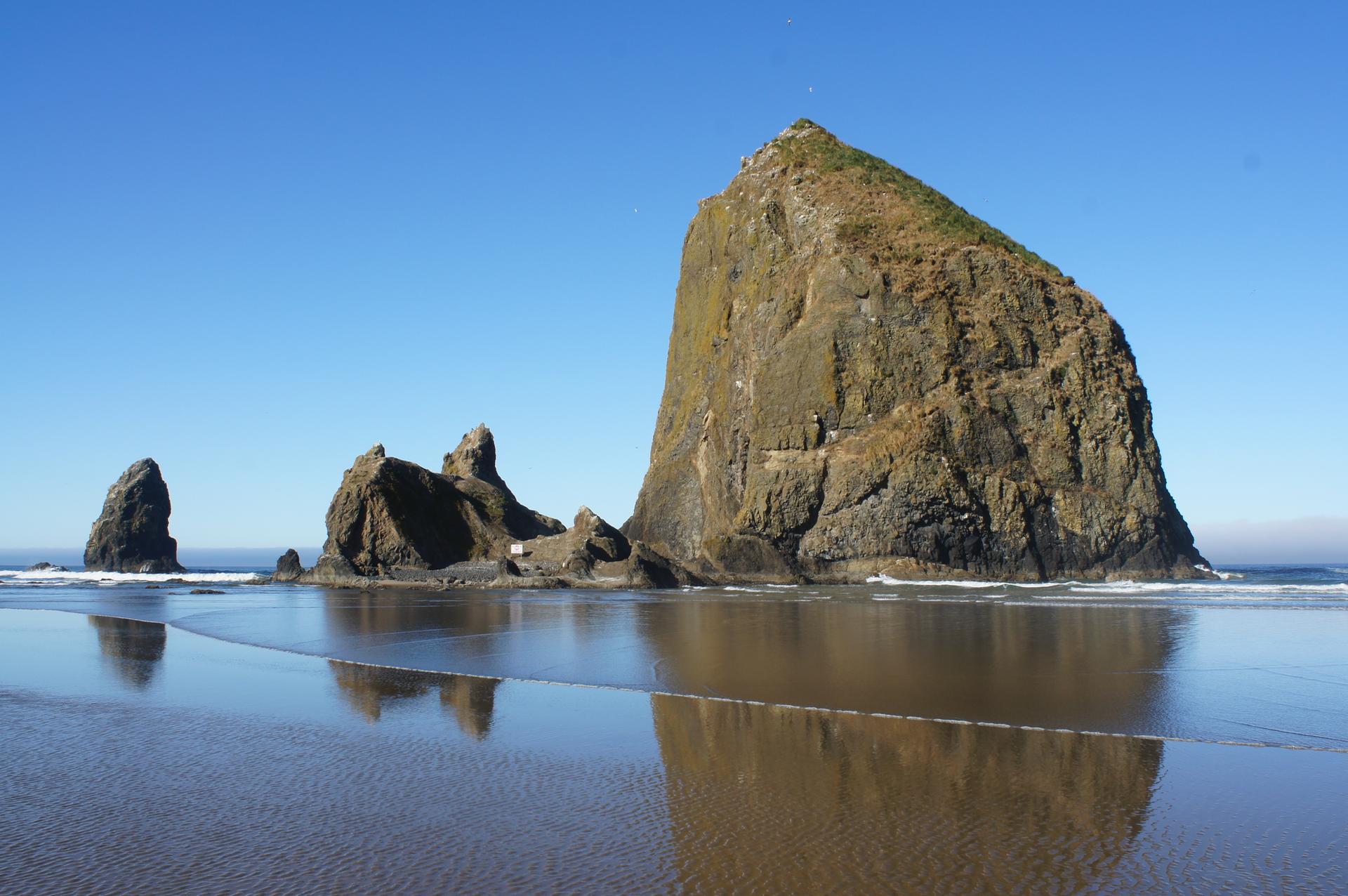 Large sea stack and smaller rocks reflected on wet beach under clear sky.