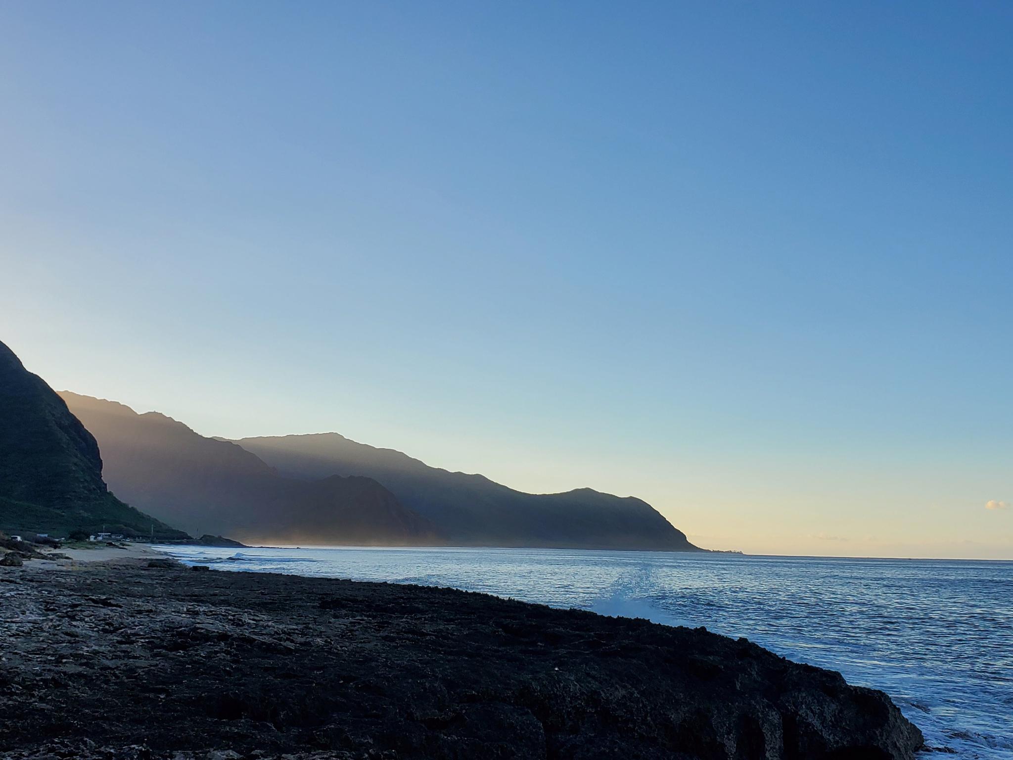 Silhouetted mountains along a coastline at sunrise with calm ocean.
