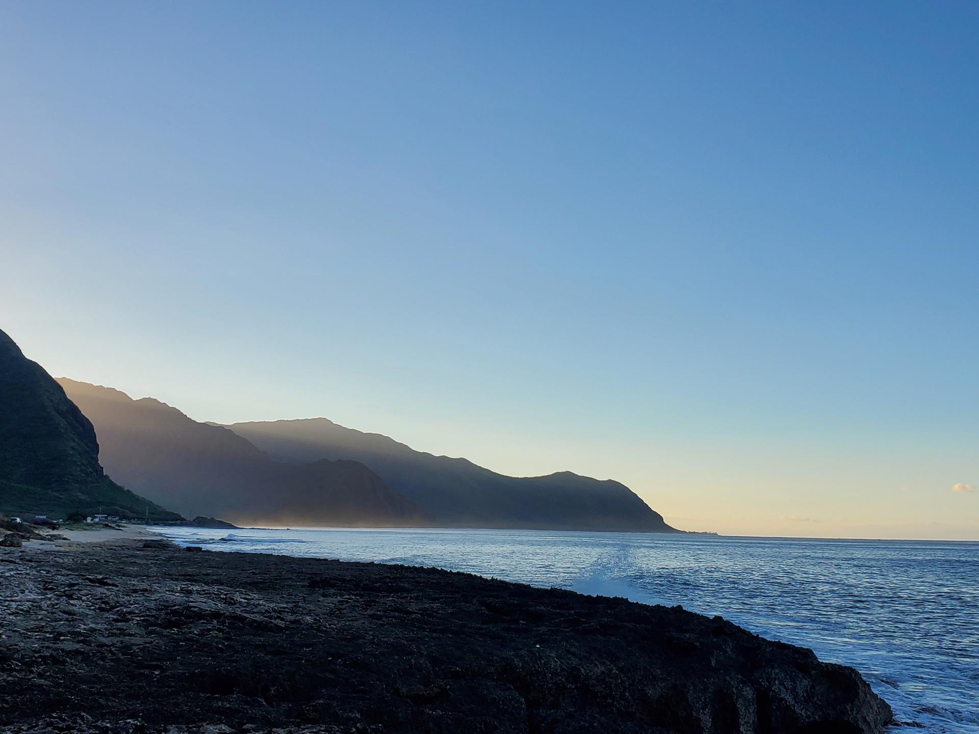 Silhouetted mountains along a coastline at sunrise with calm ocean.