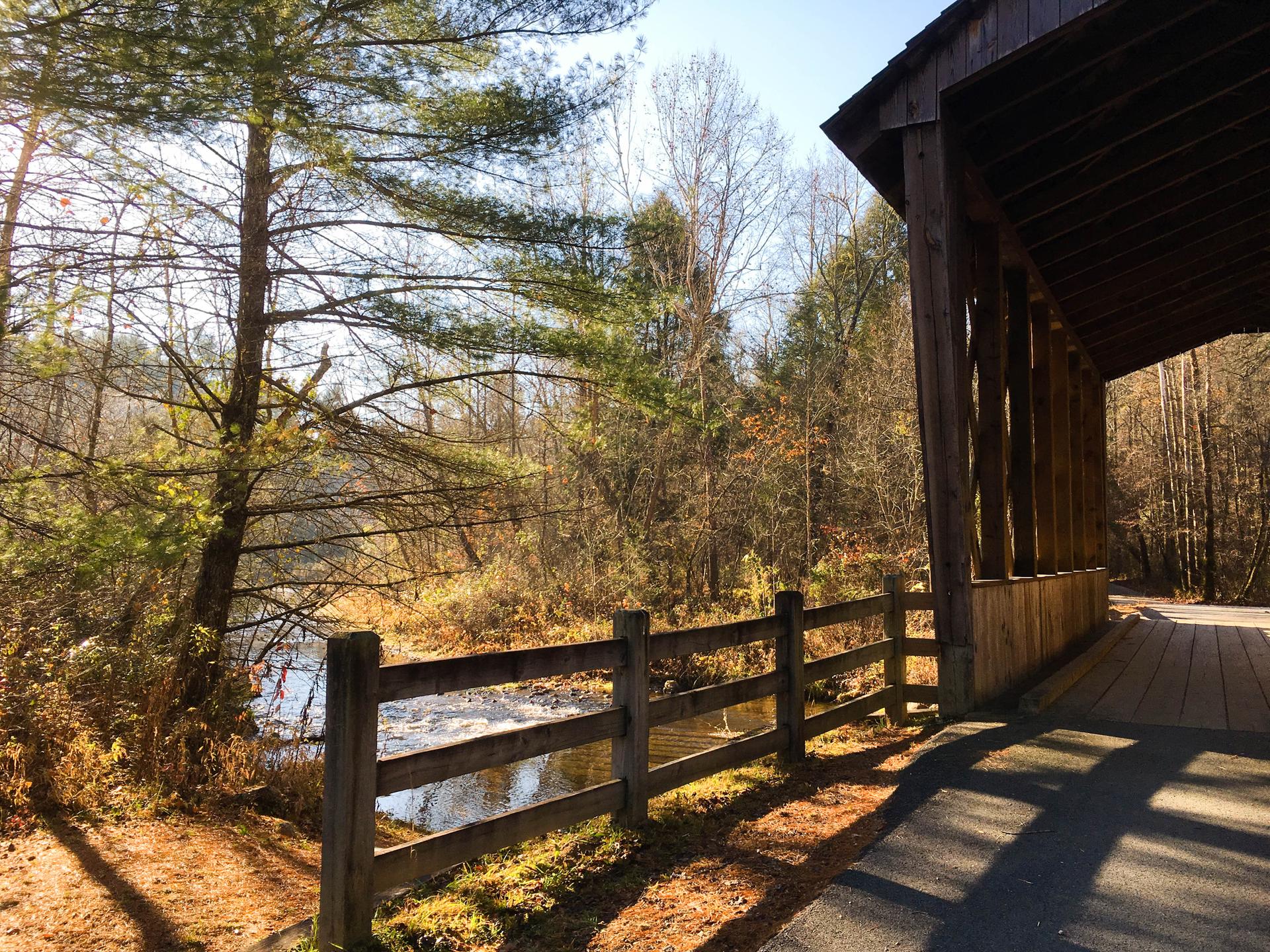 Sunny view of a wooden covered bridge next to a river and trees.