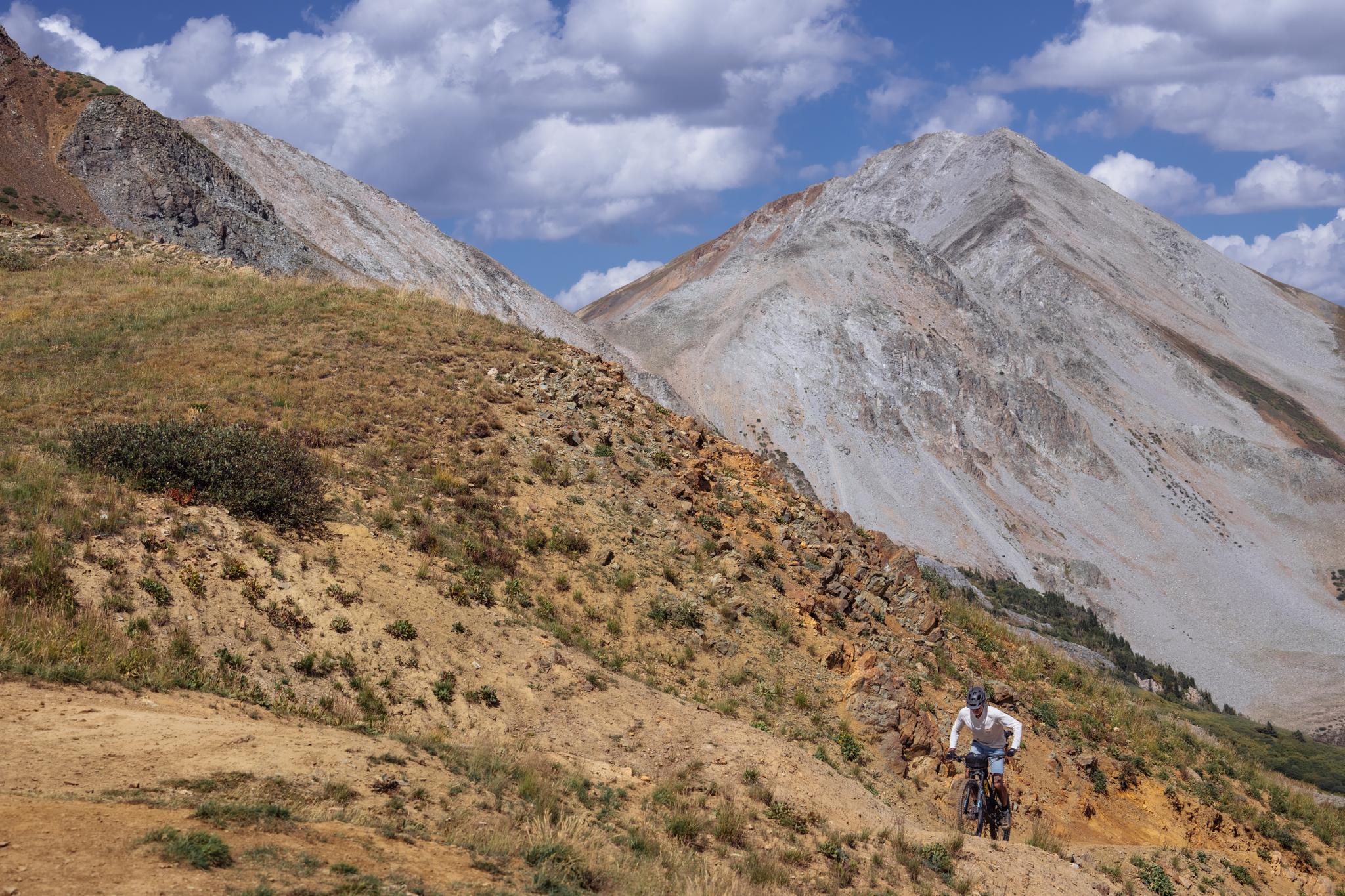Mountain biker on a rocky trail with large, arid mountains under cloudy sky.