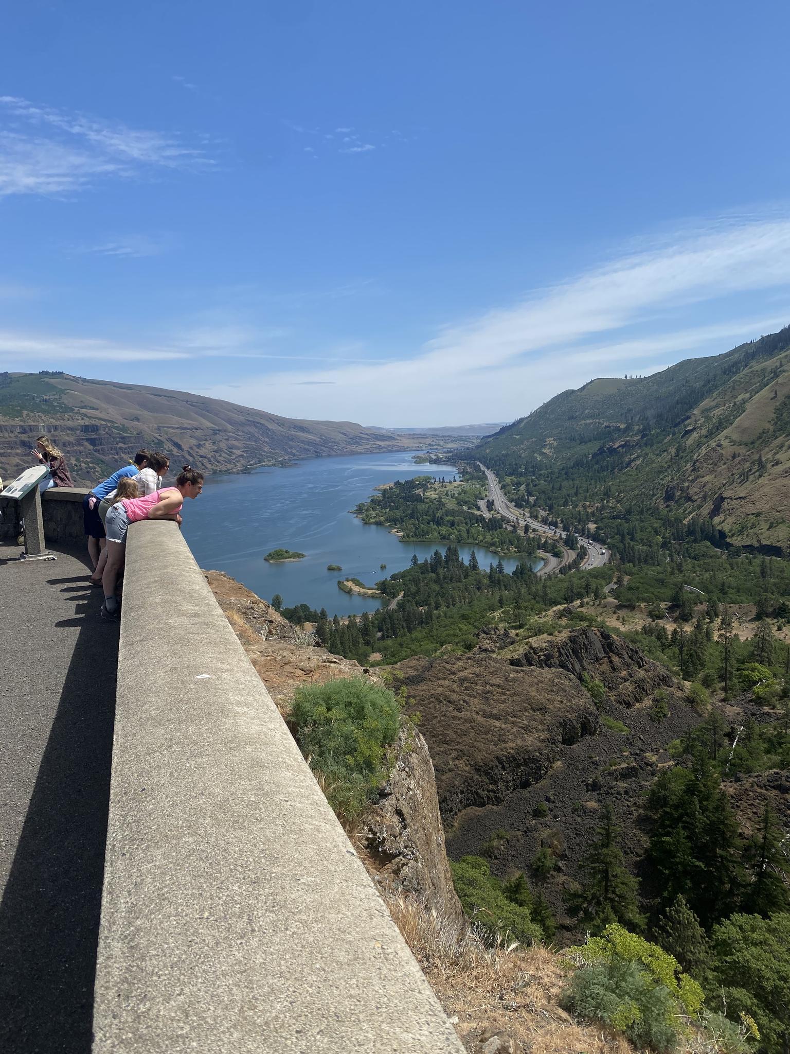 People lean over a concrete barrier, viewing a wide river valley below.