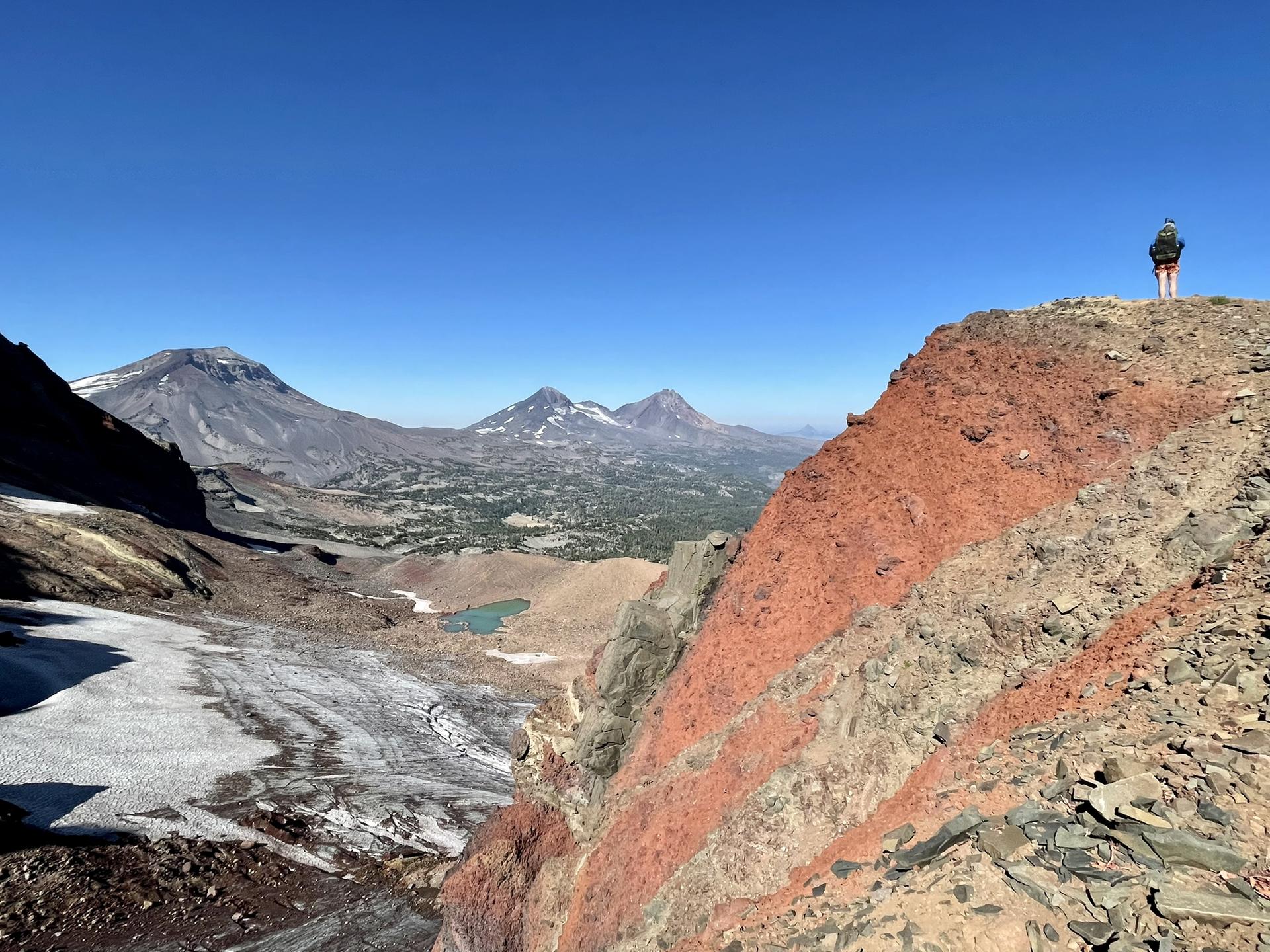 Hiker on a red rocky ridge overlooking a vast mountain landscape.