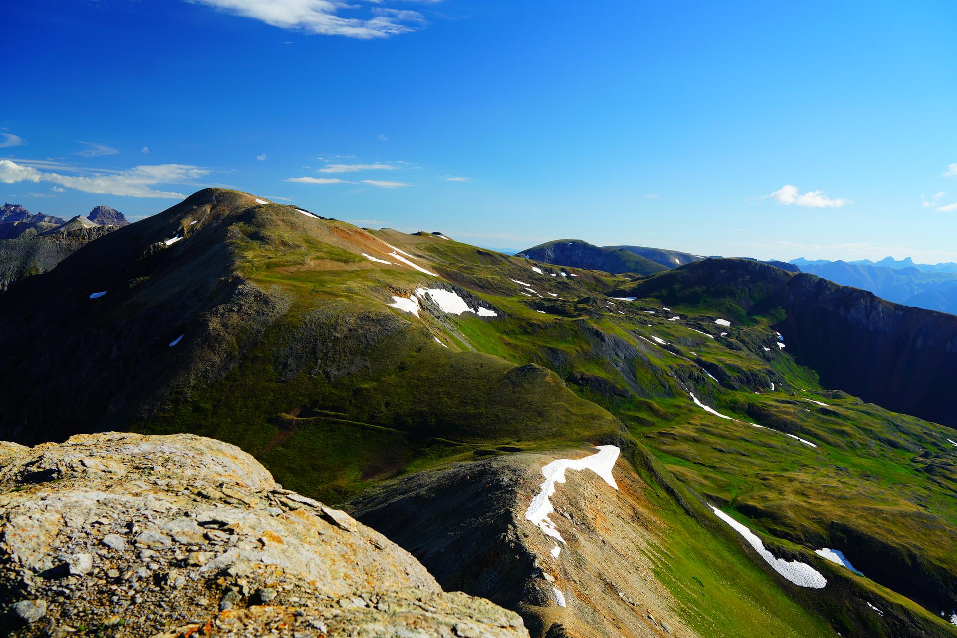 Green mountain ridge with snow patches and rocky foreground under a clear blue sky.