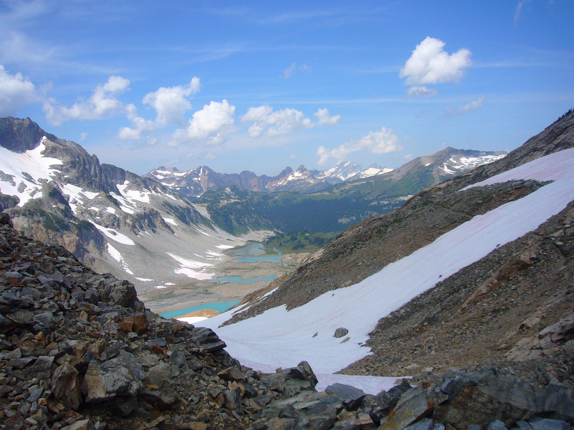 A vast mountain valley with snow-covered slopes, rocky peaks, and distant blue lakes.