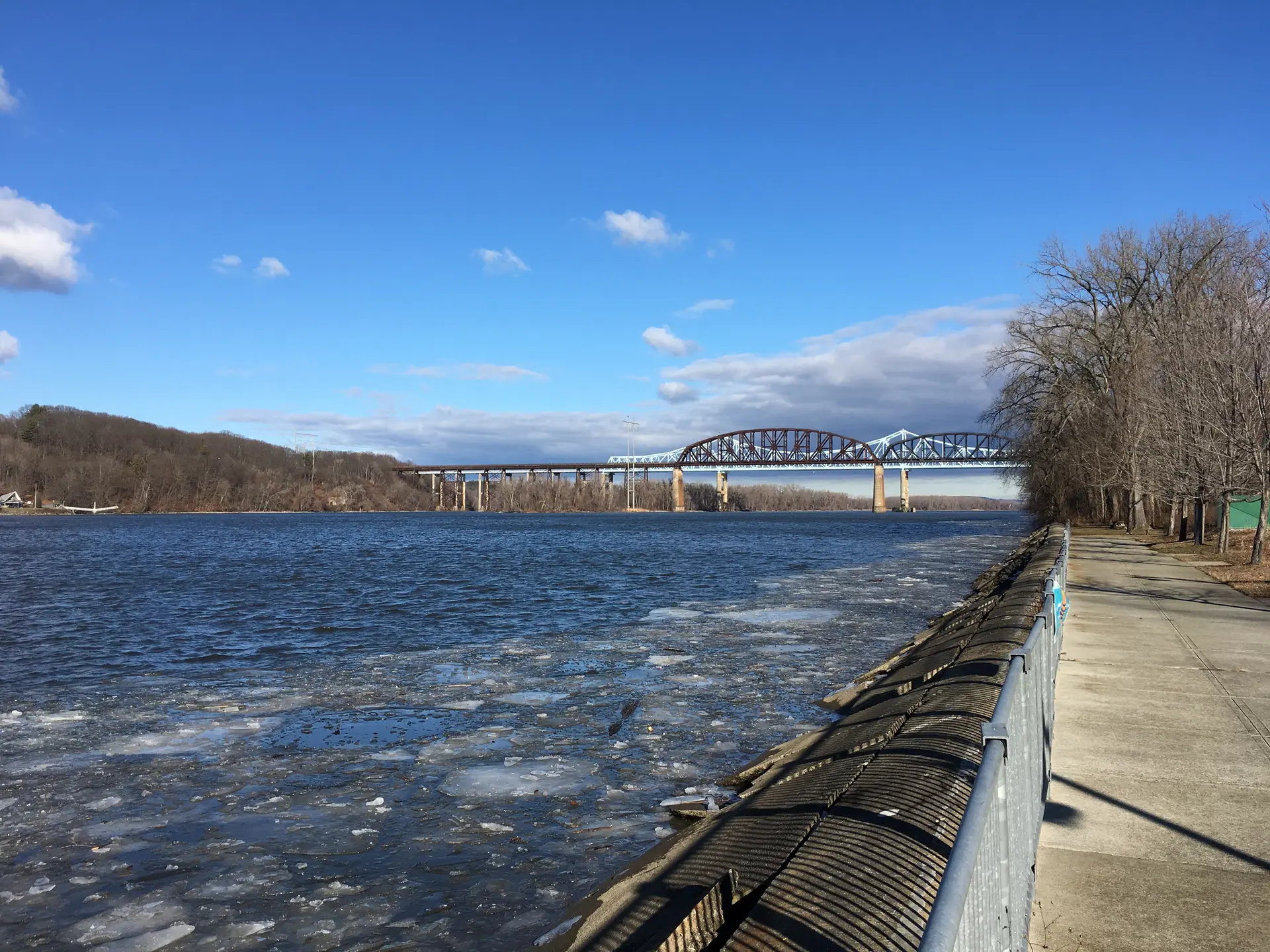 Icy river with a distant bridge, paved path, and bright blue sky.
