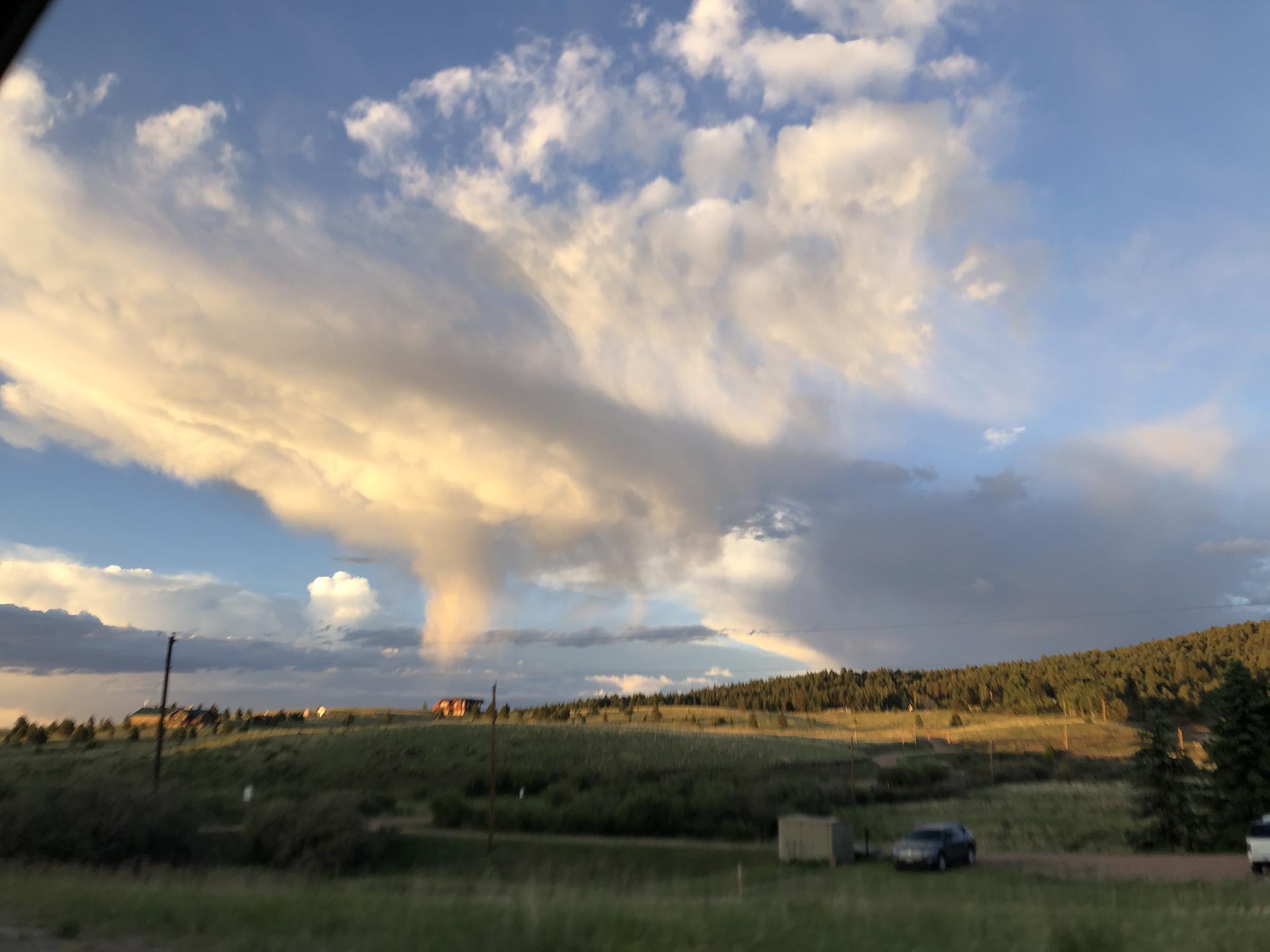 Dramatic funnel-shaped clouds over a hilly landscape with trees and a parked car at sunset.