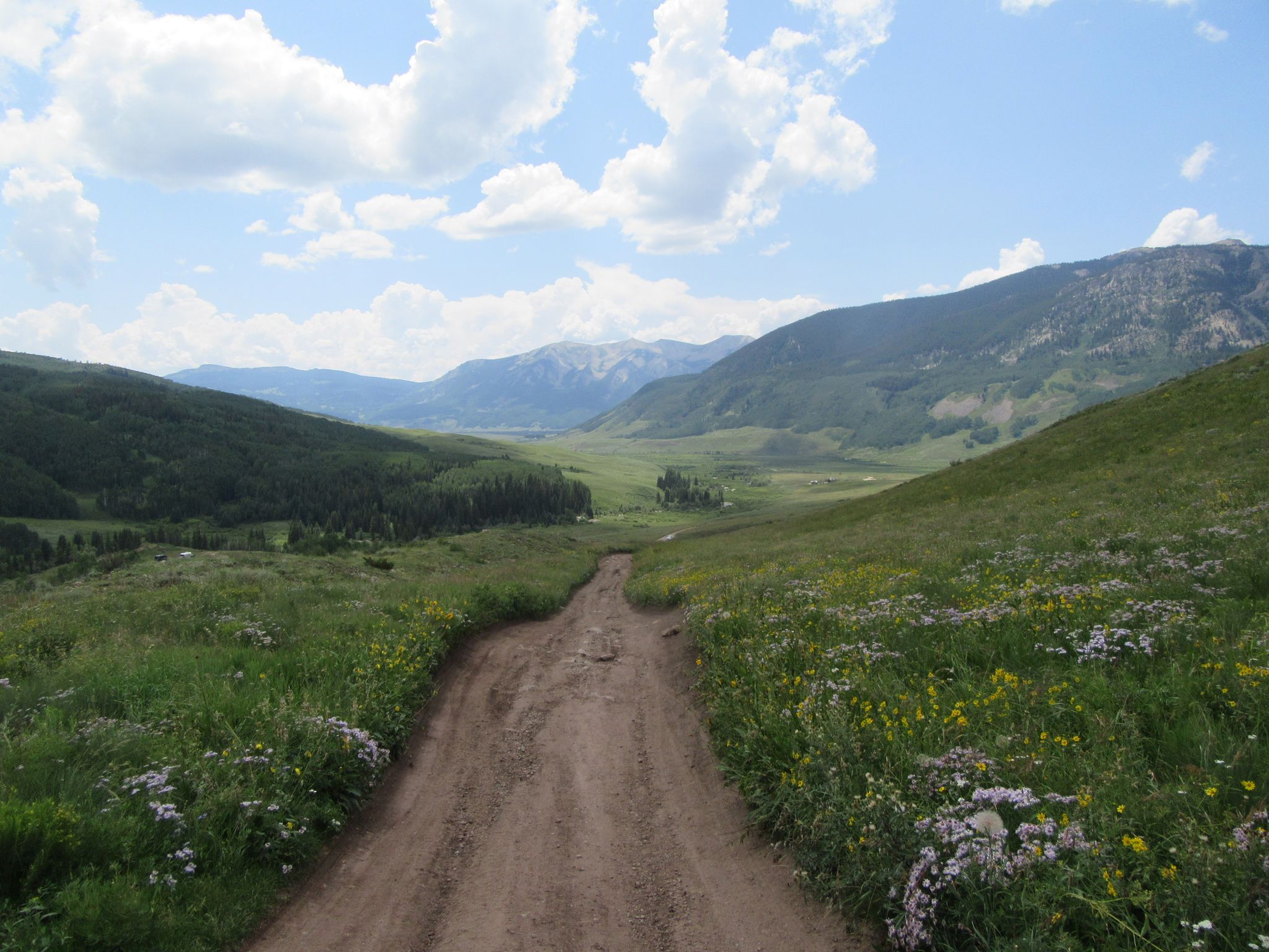 A wide dirt road descends into a lush green mountain valley under a cloudy sky. - Teocalli Ridge Loop