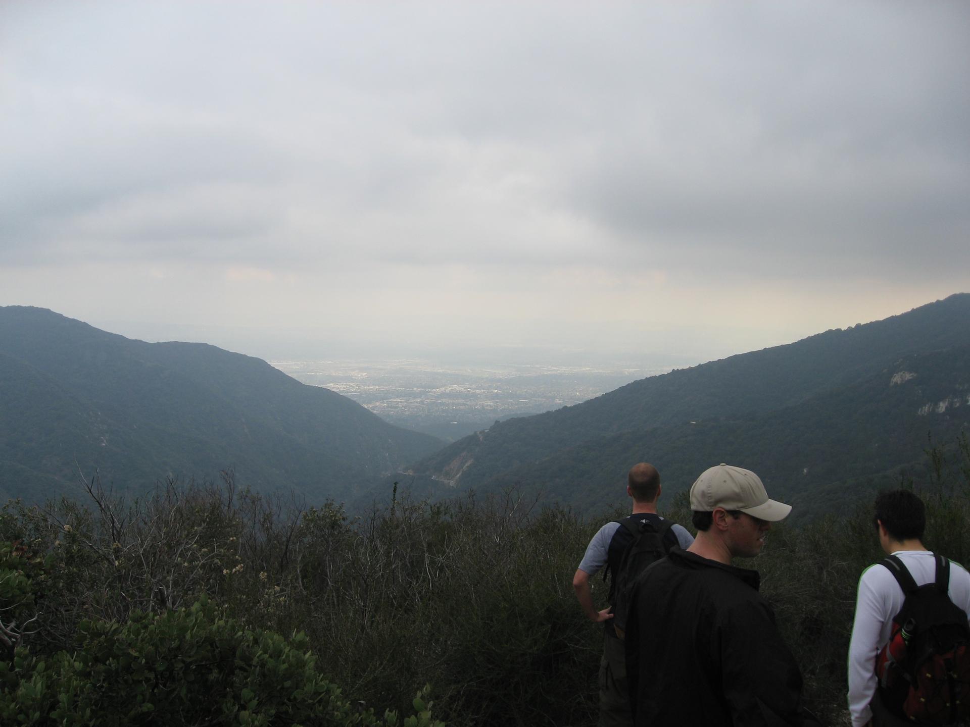 Three hikers view a distant valley and city under an overcast sky.