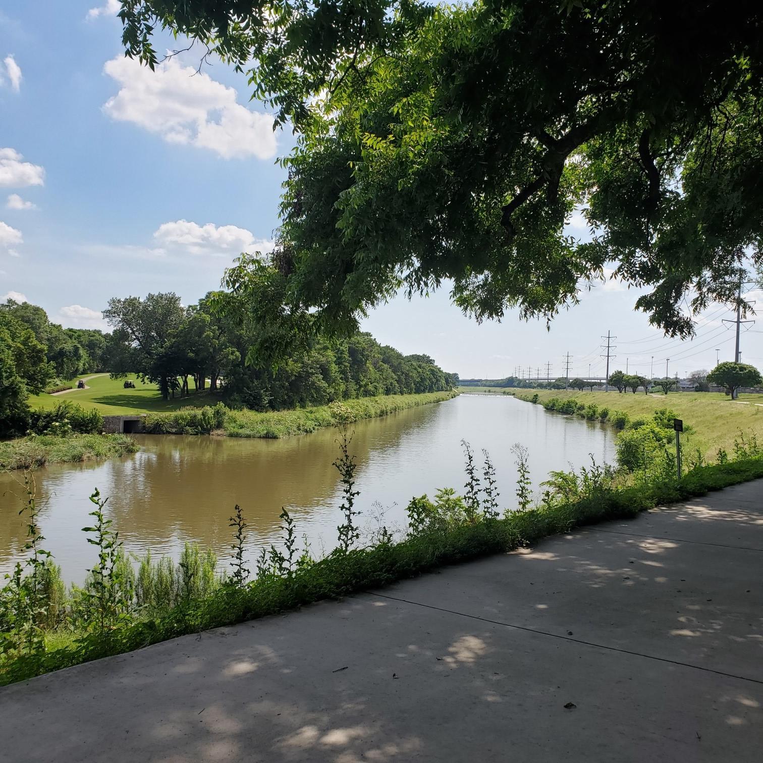 Paved path beside a wide river, shaded by lush green trees.