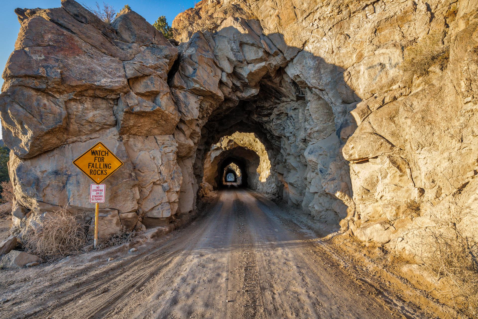 A dirt road enters a sunlit rock tunnel with a falling rocks sign.