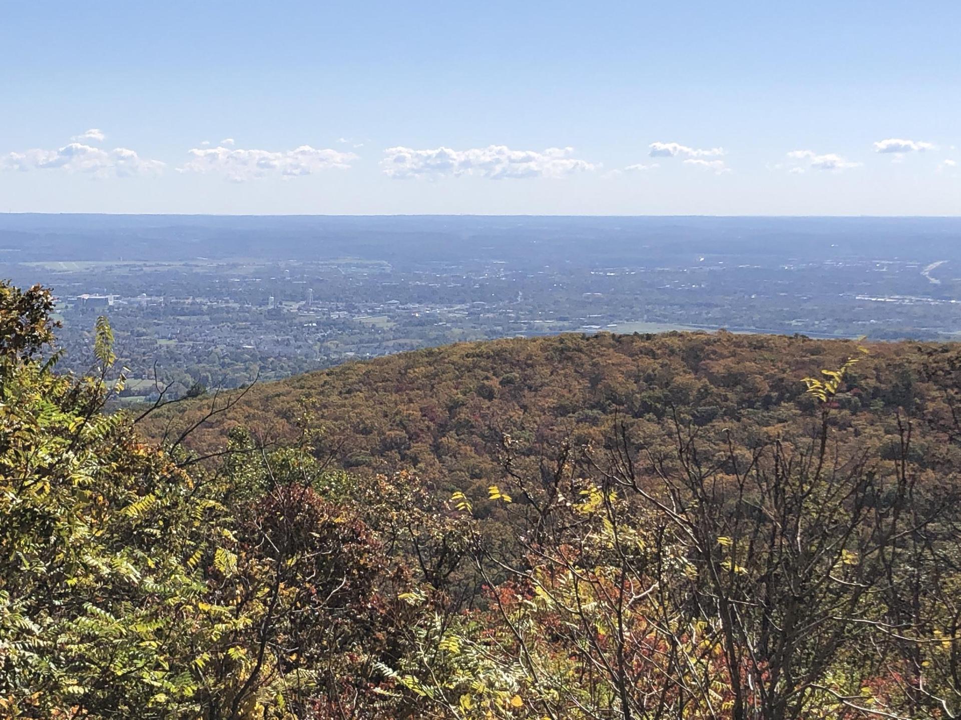 Expansive view of forested hills and a distant city under a blue sky.