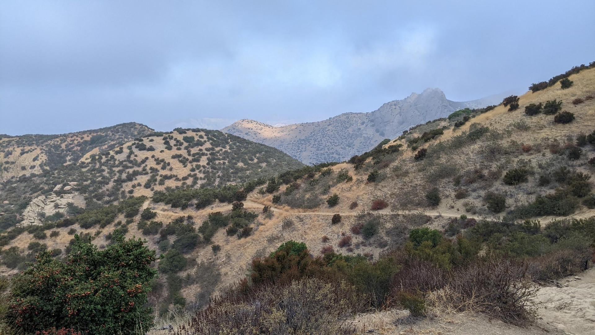 Arid mountains with a winding trail under a cloudy sky.