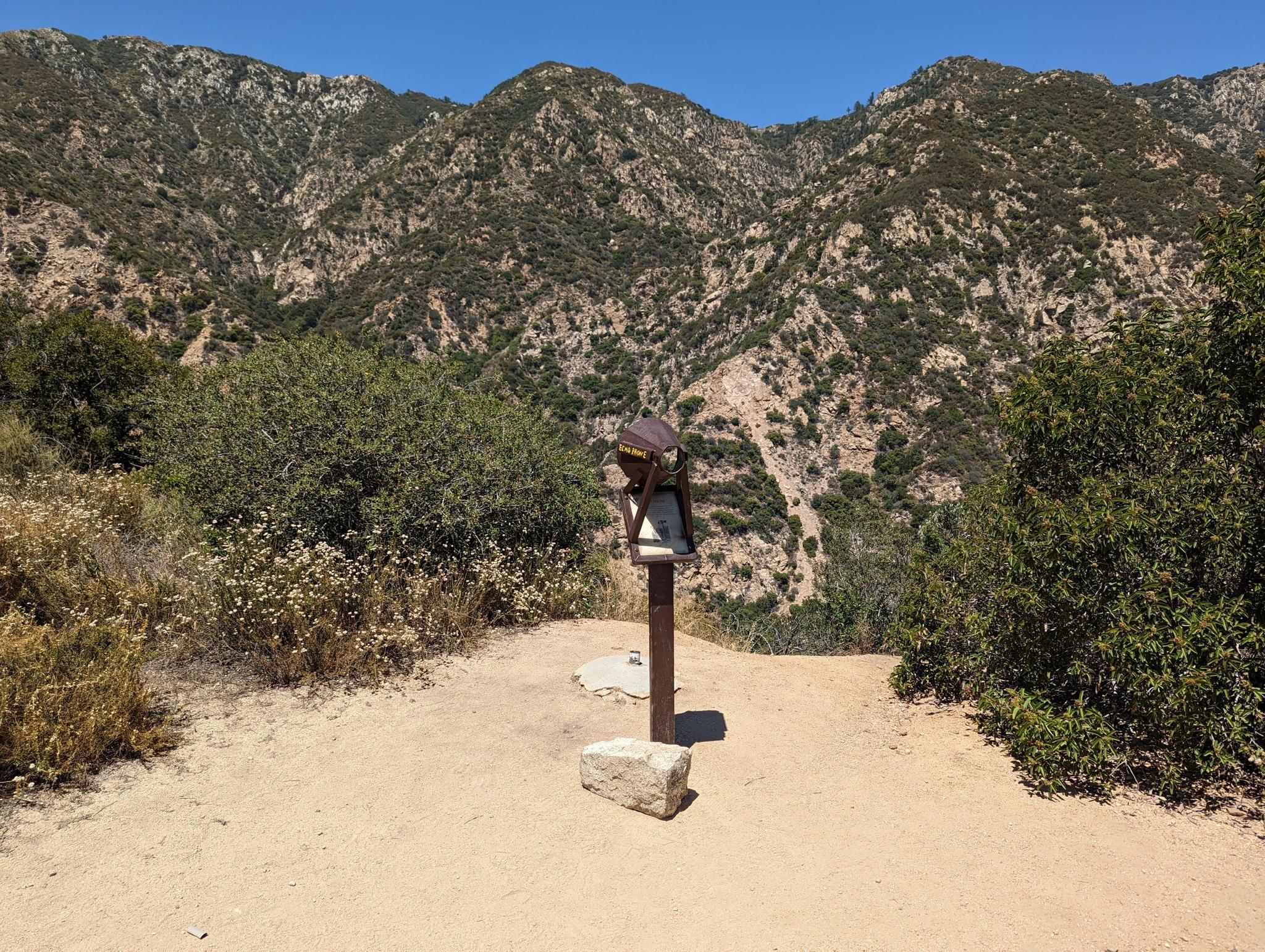 Wooden stand on a dirt path overlooking steep, brushy mountains under a clear sky.
