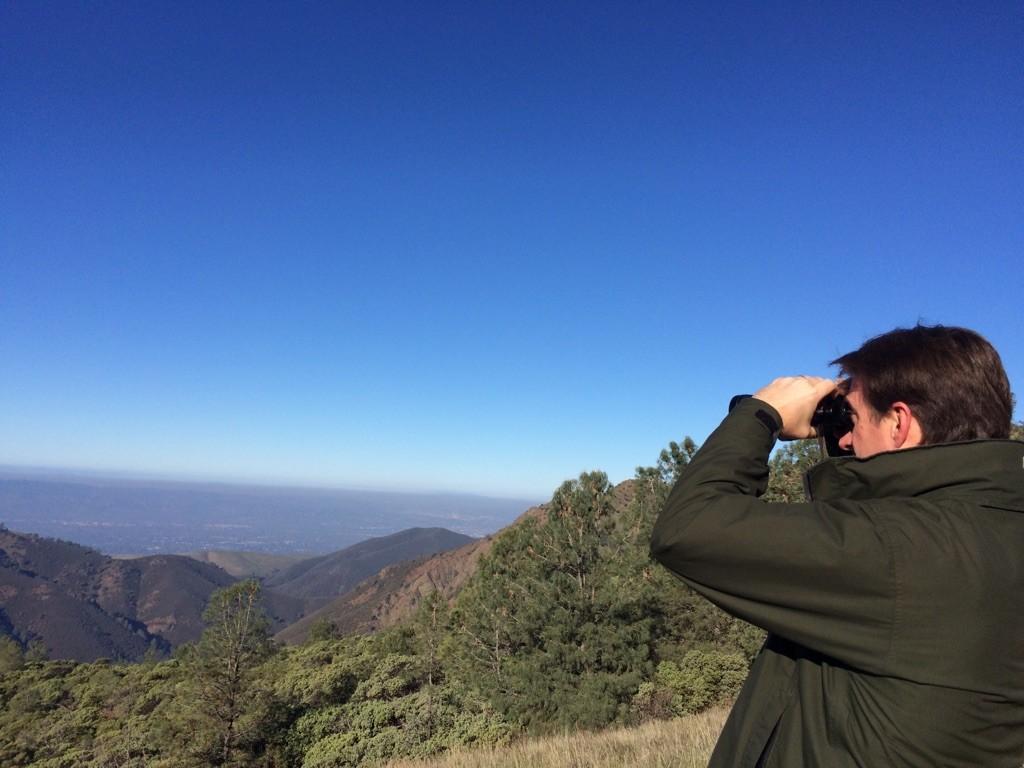 Man in green jacket views vast mountain landscape through binoculars.