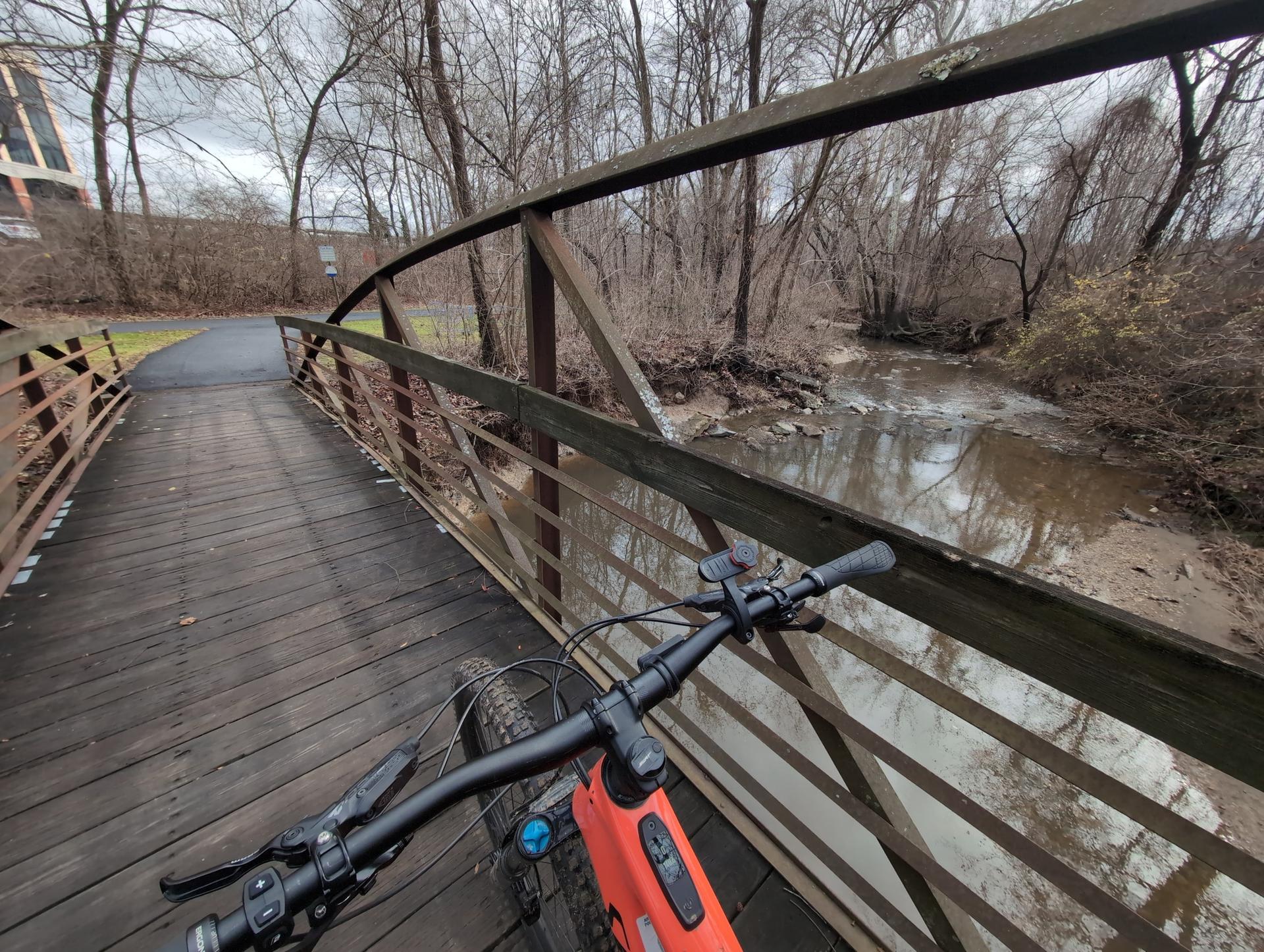Bicycle handlebars on a wooden bridge overlooking a river and bare trees.