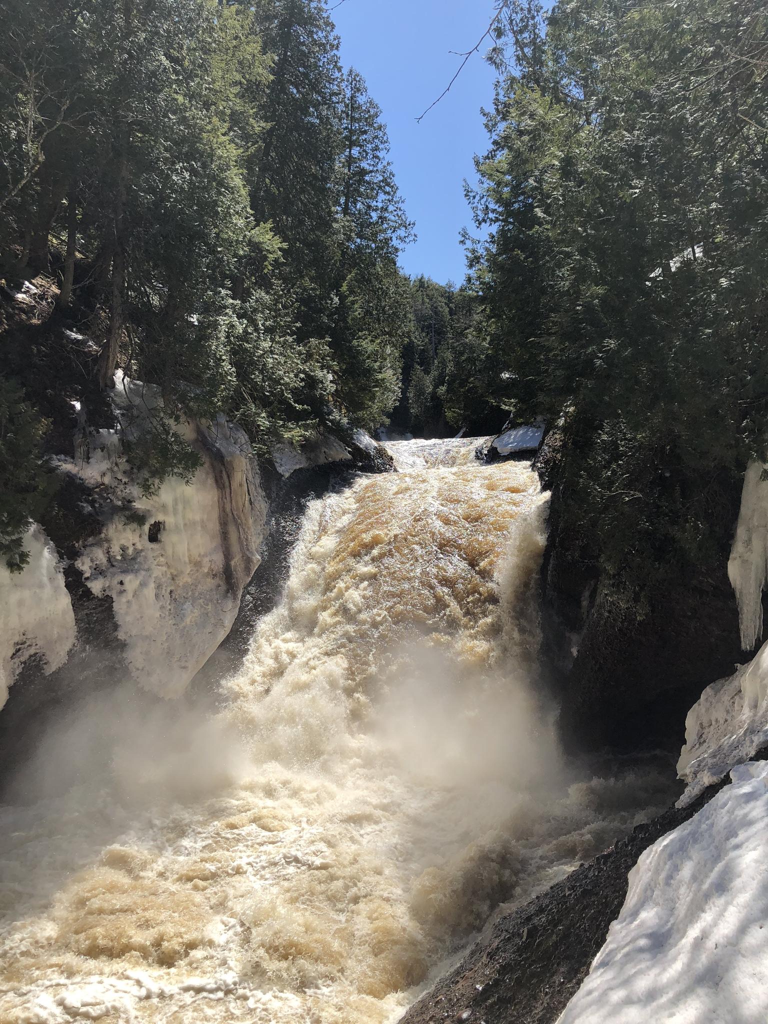 A powerful waterfall plunges between snowy, forested banks under a blue sky.