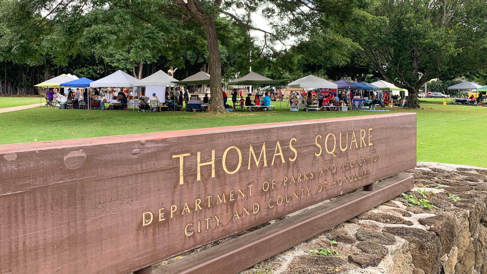 Wooden sign in foreground, outdoor market with tents in a green park.